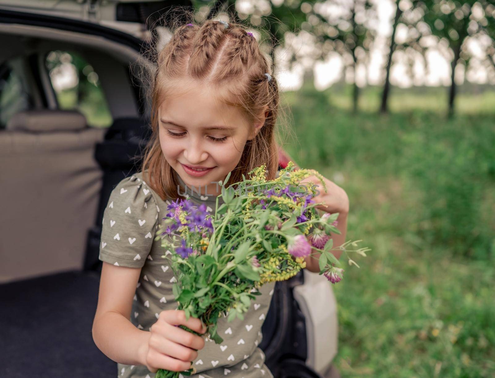Beautiful little girl with cute hairstyle sitting in the car trunk holding flower bouquet and looking down. Child kid with summer plants in the vehicle at the nature
