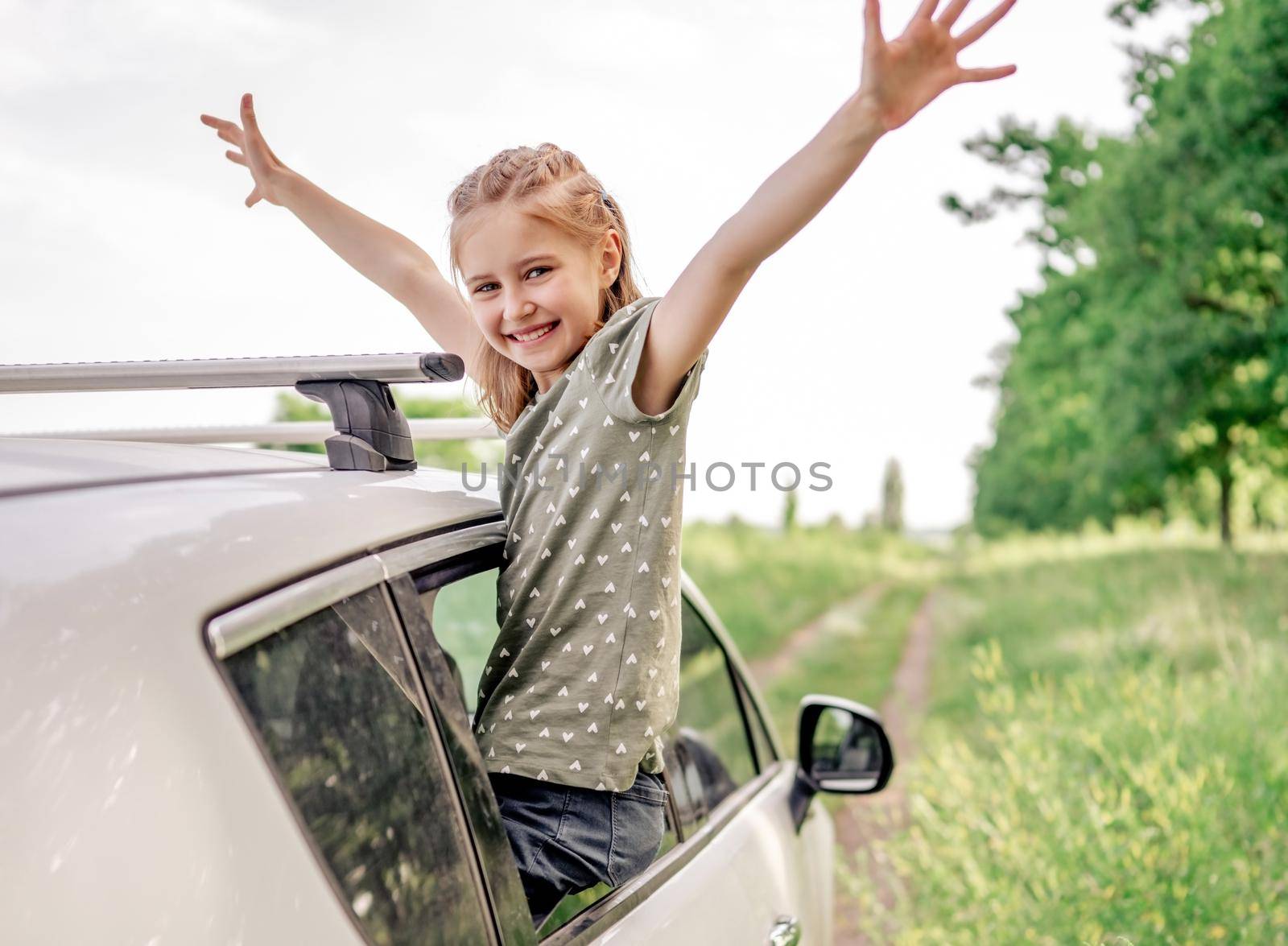 Beautiful preteen girl sitting in the car window open, smiling and holding hands up in the field. Happy child kid in the vehicle outdoors during summer journey