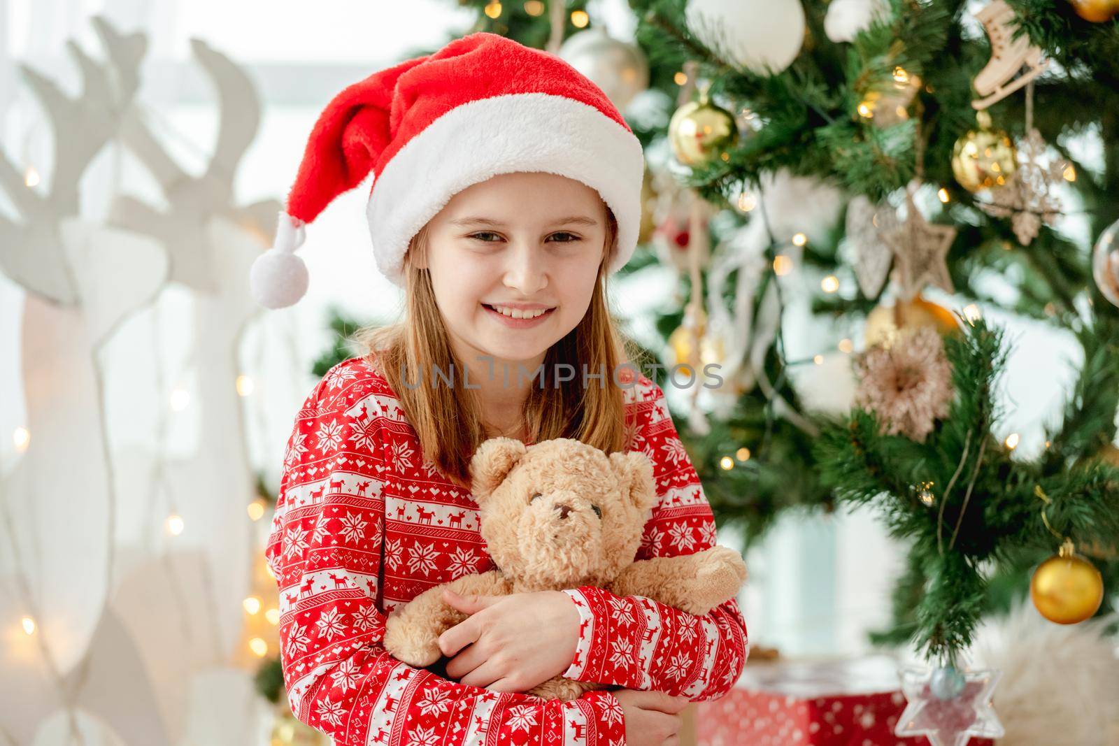 Happy child girl hugging teddy bear toy at home with Christmas tree on background. Cute kid with gifts at New Year time