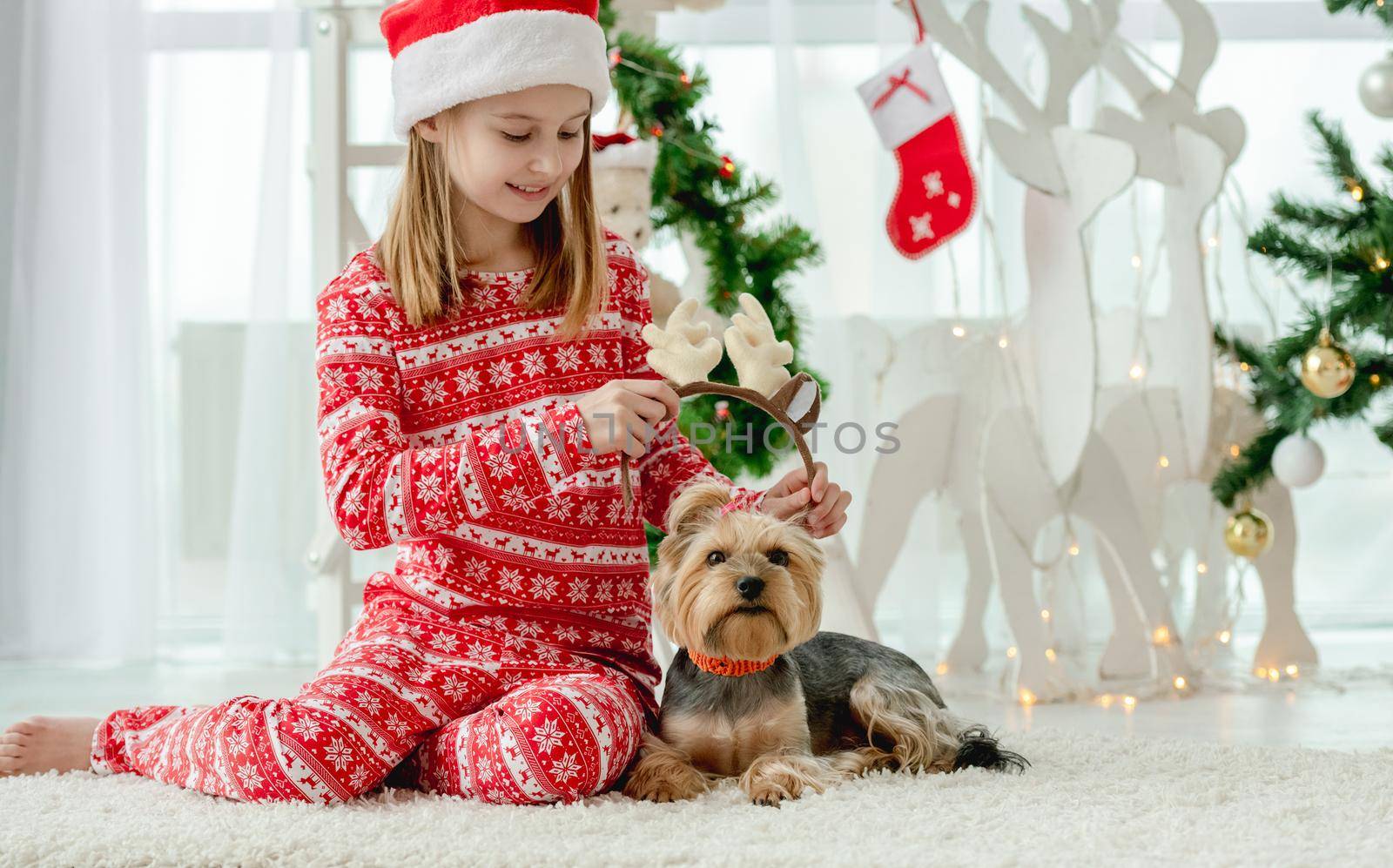 Child girl with dog at Christmas time at home with holiday decoration. Kid wearing red costume and pet doggy in room with New Year decoration