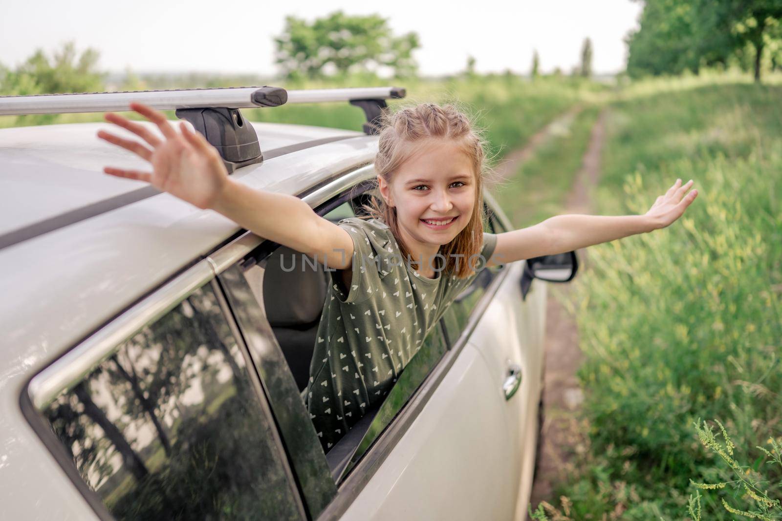 Beautiful preteen girl sitting in the car and looking out the window open, smiling and holding hands up in the field. Happy child kid in the vehicle outdoors during summer journey