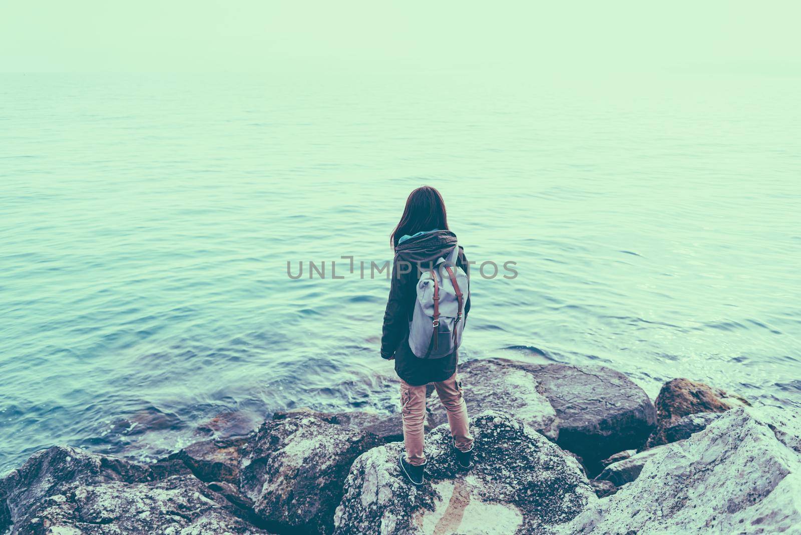 Traveler young woman standing on stone on coastline and enjoying view of sea, rear view. Image with instagram filter