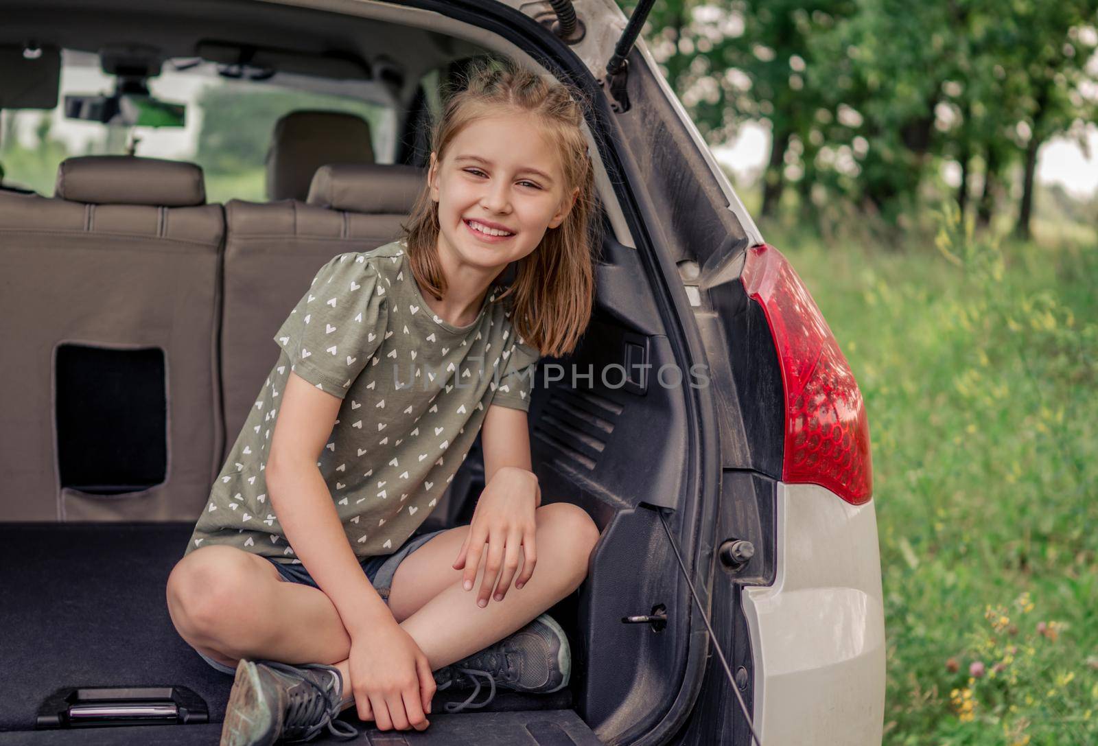 Preteen girl with beautiful hairstyle sitting in the car trunk and smiling looking at the camera. Happy child kid in the vehicle at the nature during summer trip