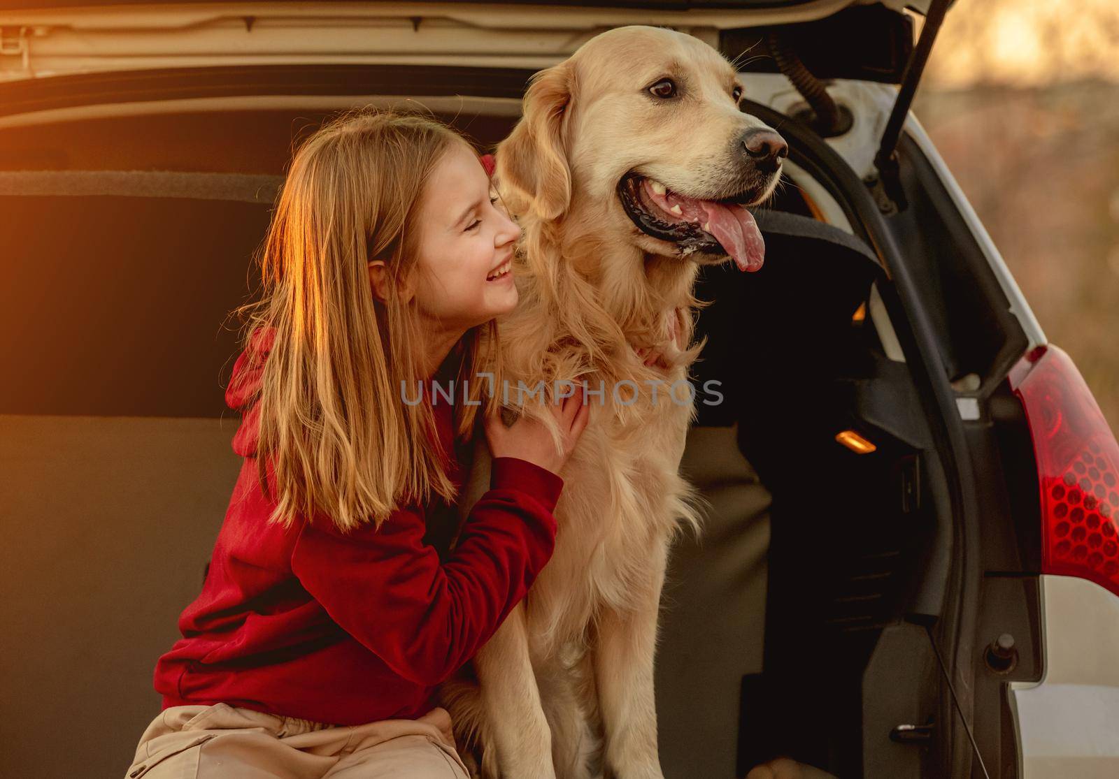 Girl child hugging golden retriever dog sitting in car trunk outdoors. Pretty kid petting doggy pet labrador and smiling in vehicle at nature