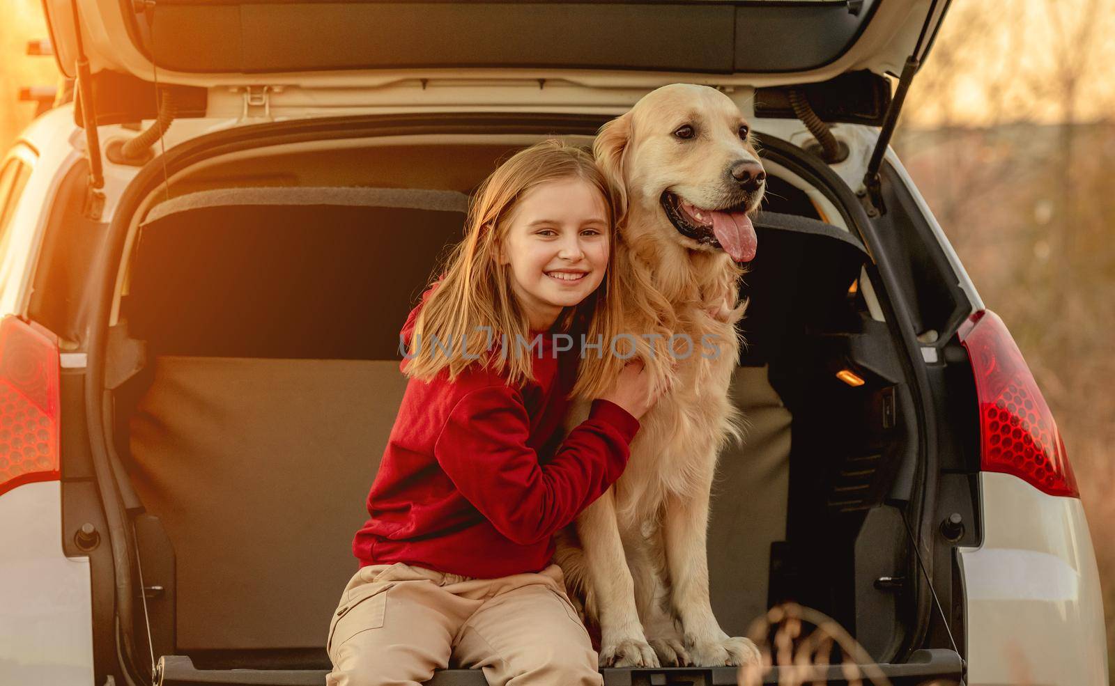 Girl child hugging golden retriever dog sitting in car trunk outdoors. Pretty kid petting doggy pet labrador and smiling in vehicle at nature