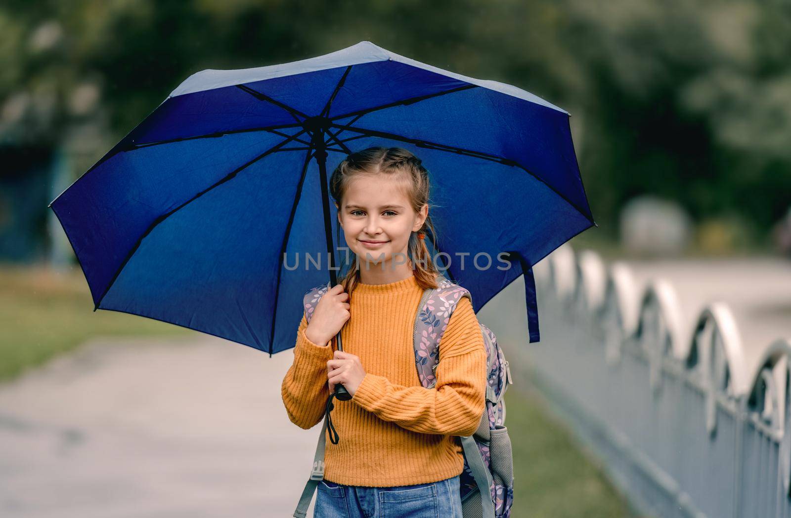 School girl with backpack and umbrella outdoors. Pretty child kid walking in rainy weather