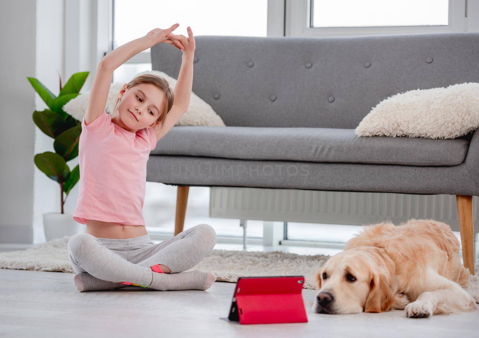Little girl stretch her back with online yoga lessons on red tablet and sitting on the floor with golden retriever dog