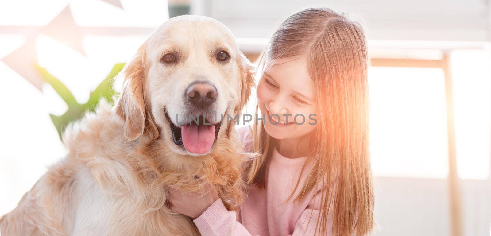 Little beautiful girl petting adorable golden retriever dog in the sunny room at home
