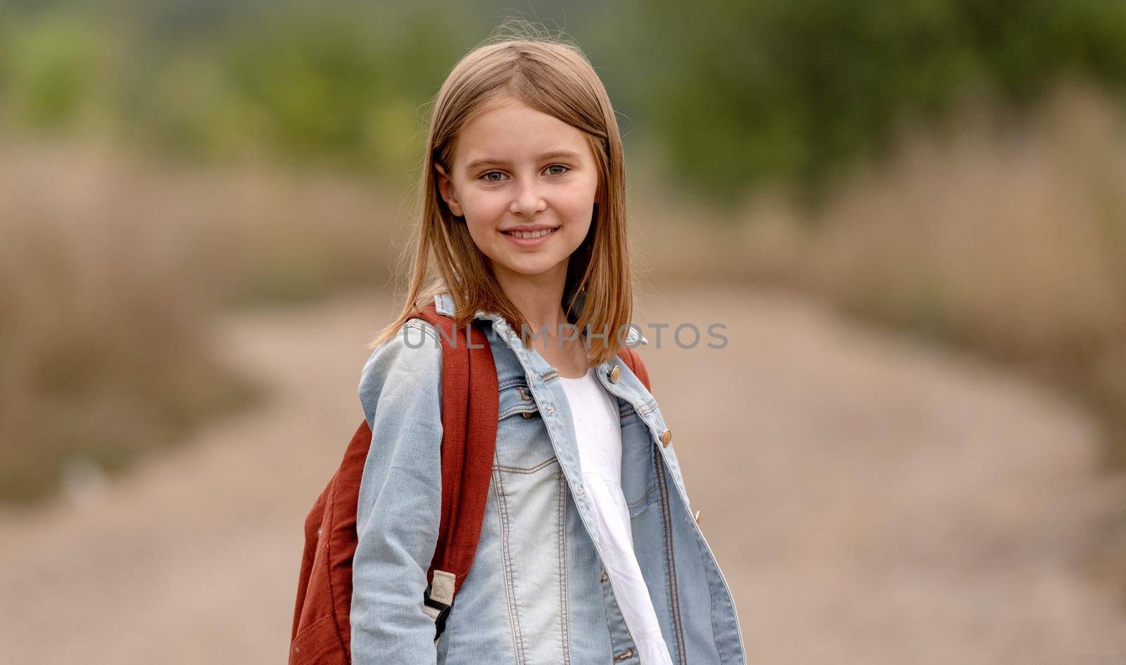 School girl with backpack looking at camera in the field with sunlight. Portrait of preteen child kid at nature