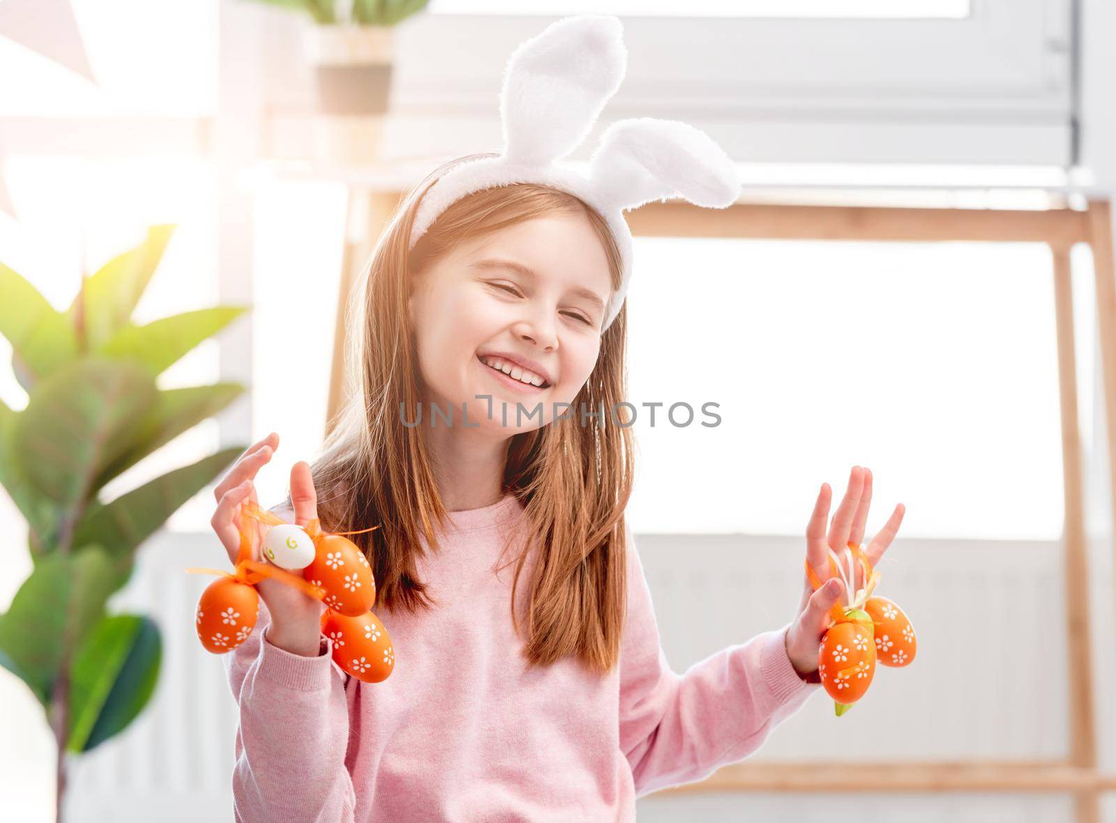 Beautiful little girl holding painted eggs in her hands and smiling at Easter day. Cute child wearing bunny ears