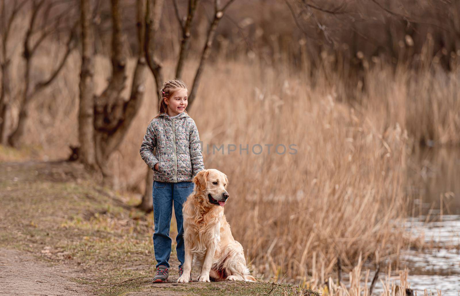 Preteen beautiful girl standing with golden retriever dog on the nature and looking at the river