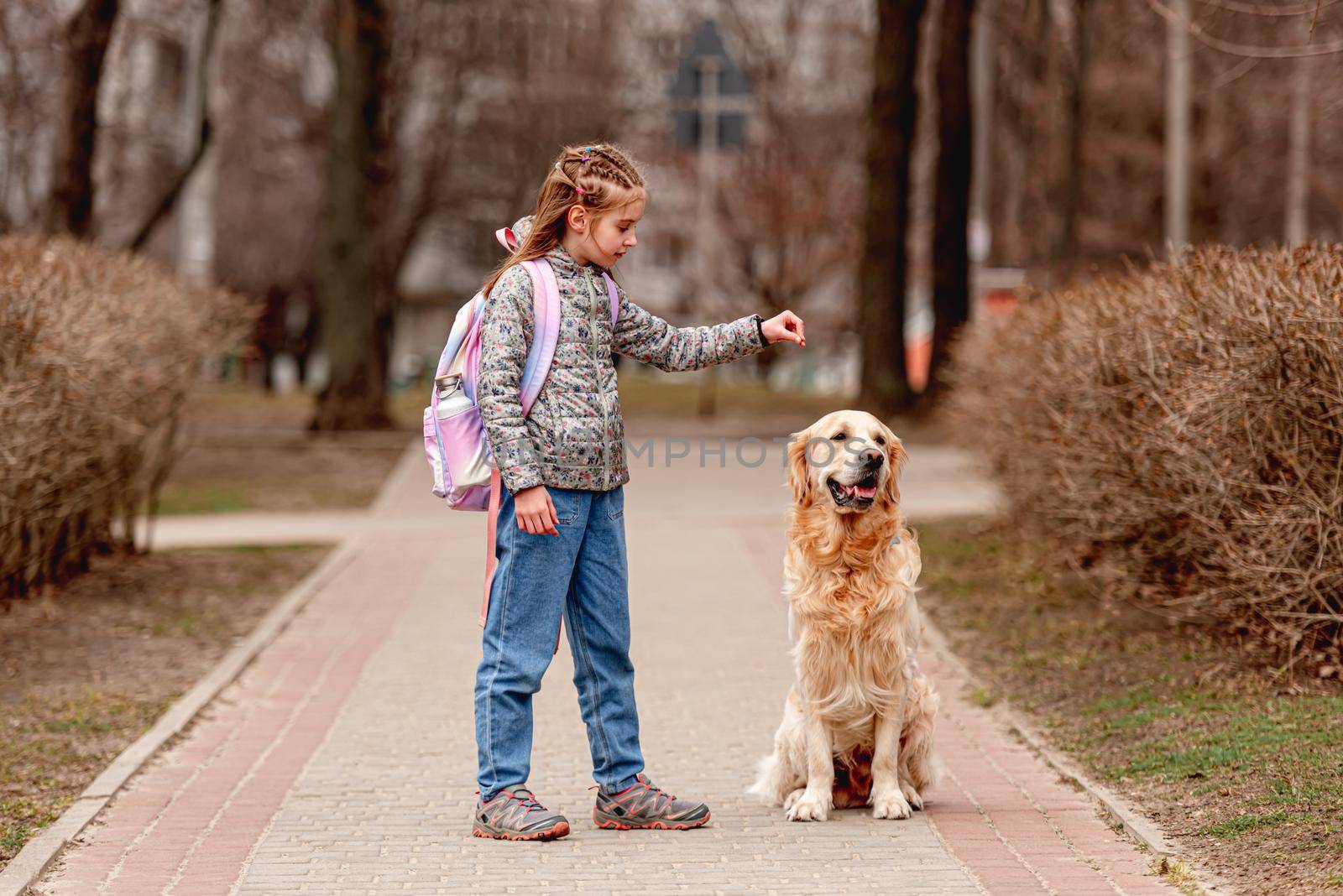 Little beautiful girl with backpack training golden retriever dog on the nature