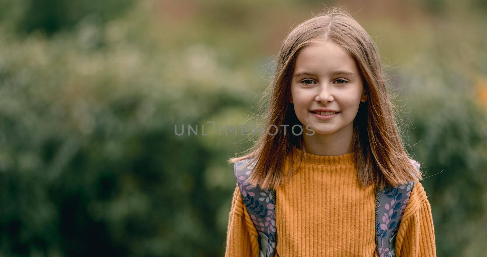 Cute school girl with packpack in the park portrait. Pretty female kid with beautiful eyes looking at camera and smiling outdoors