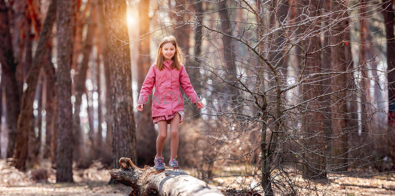 Little girl wearing pink coat walks on a log in the wood in beautiful sunny spring day