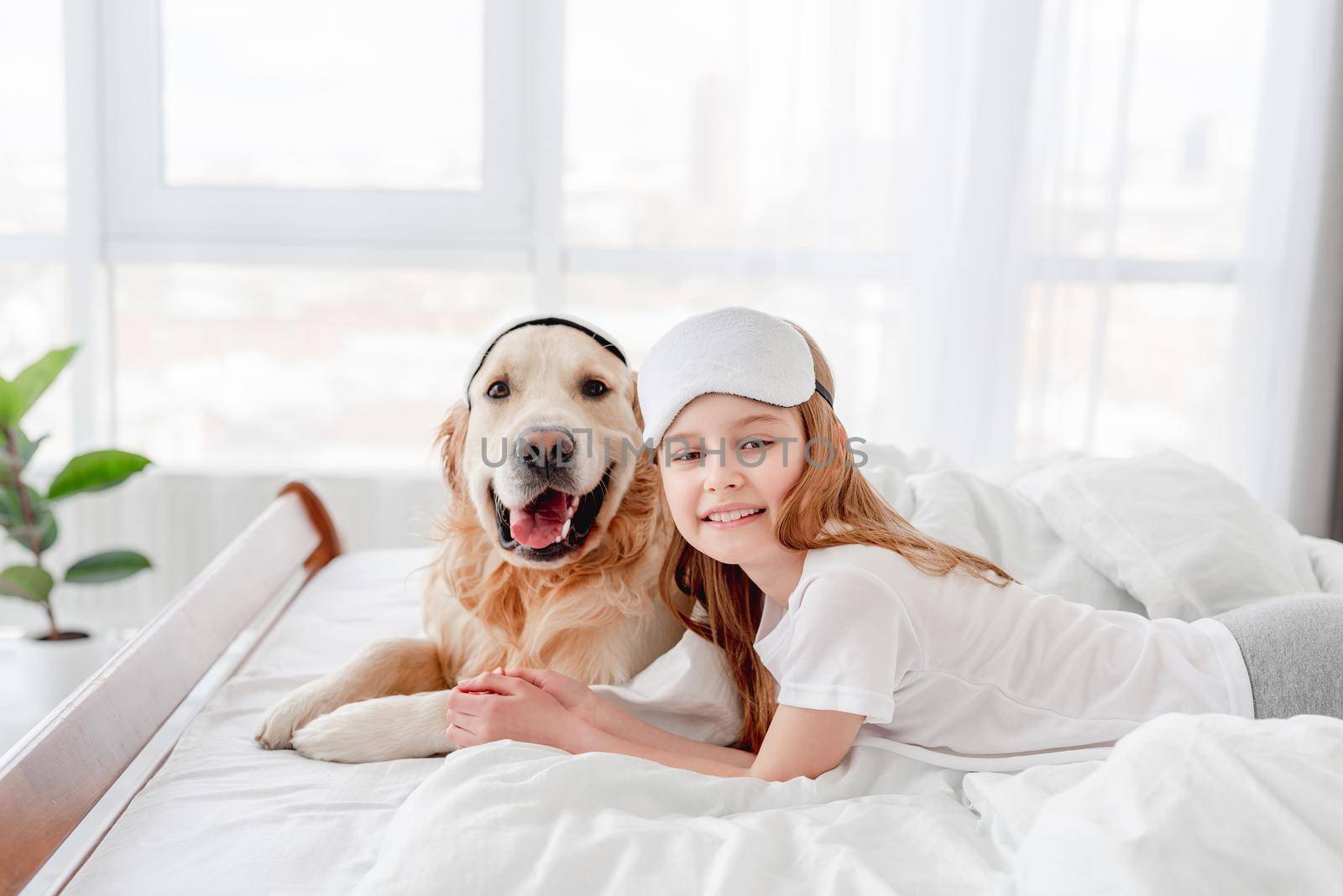 Little smiling girl and golden retriever dog wearing eye sleeping mask staying in the bed together and looking at the camera. Portrait of kid with pet in the morning time. Doggy with owner at home
