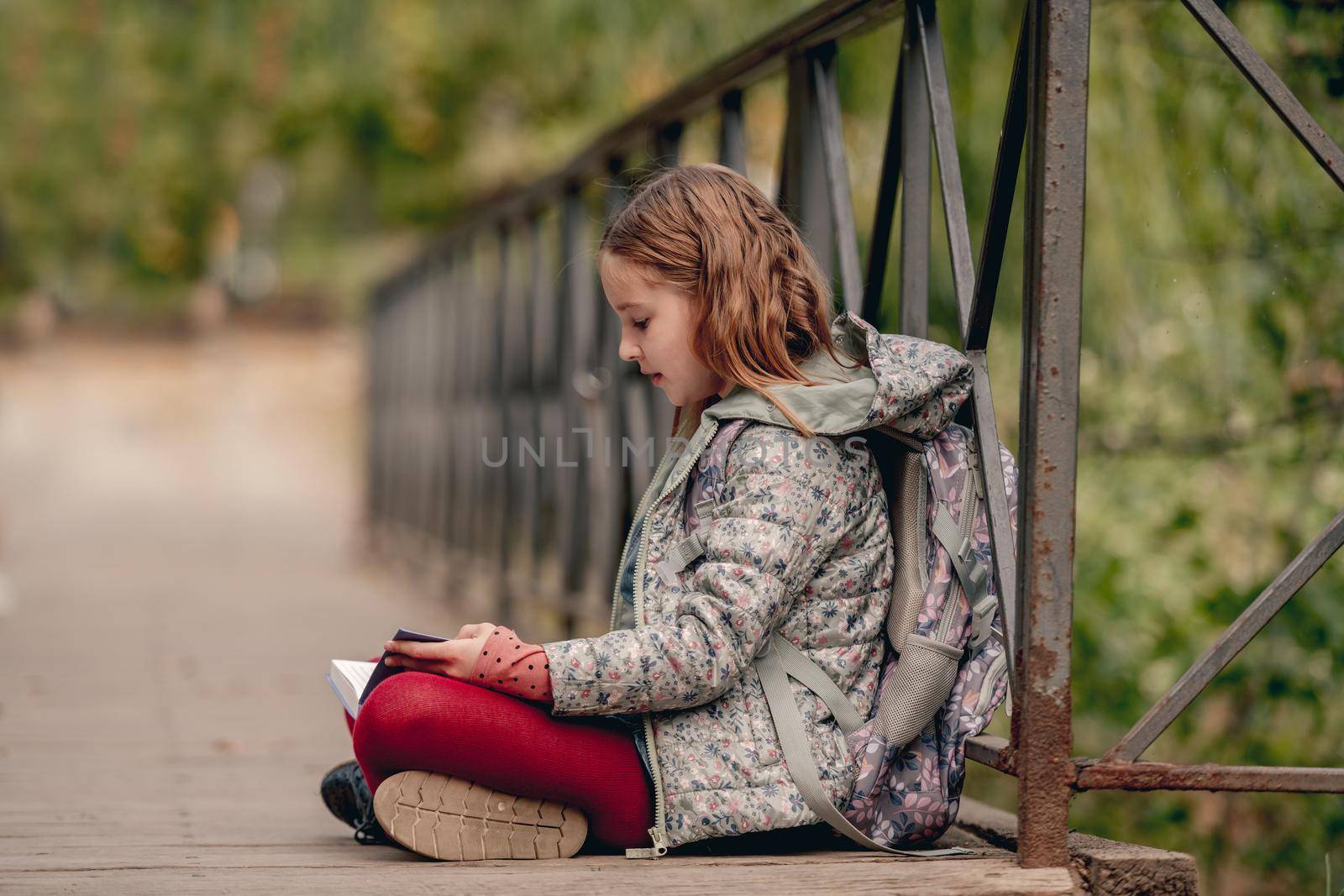 Preteen school girl with backpack sitting on ground and reading book outdoors. Sweet pupil female kid studying in autumn park