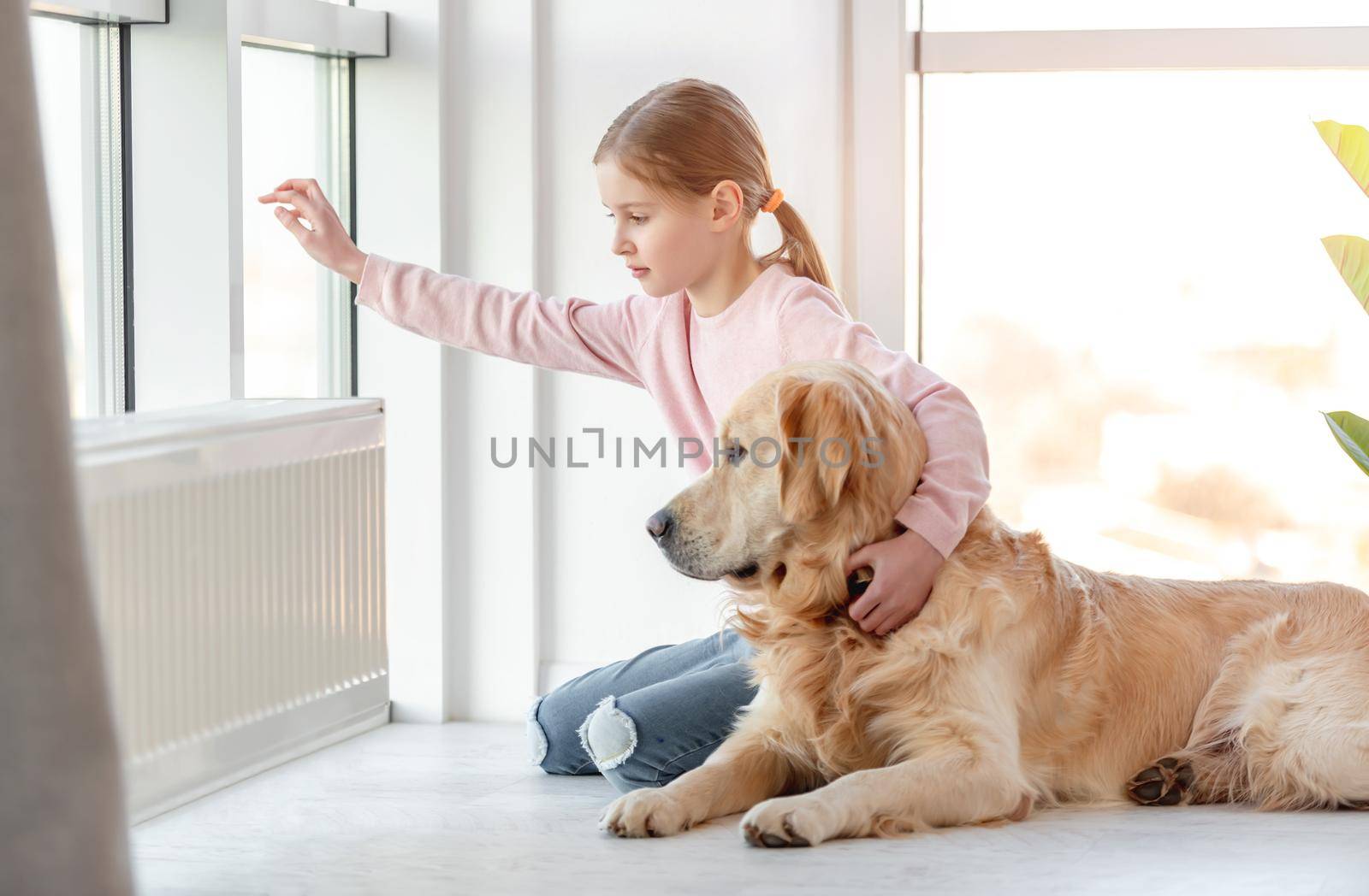 Little girl child with golden retriever dog sitting on the floor and looking out the window