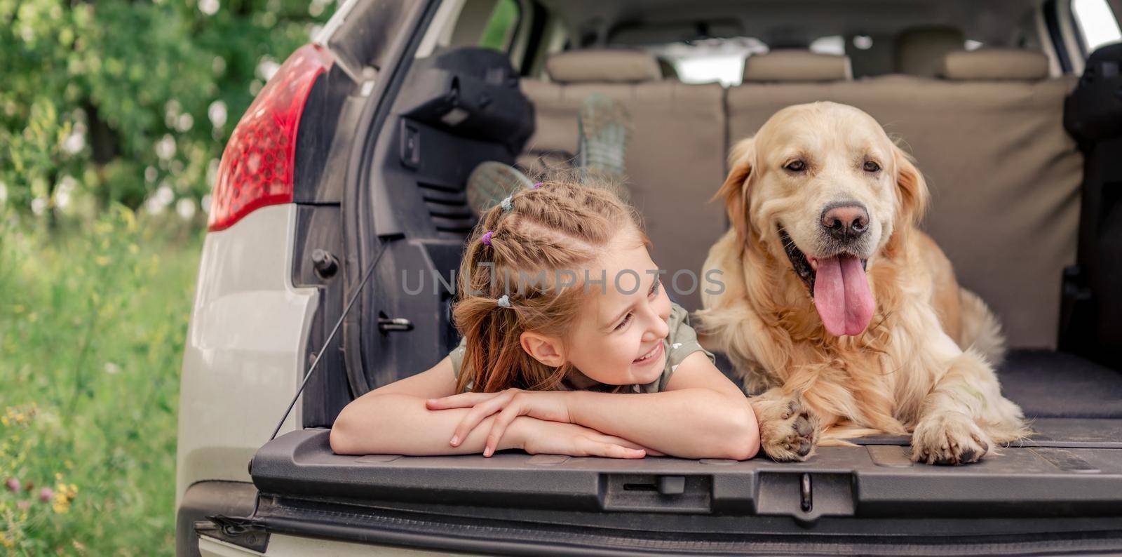 Beautiful little girl lying with golden retriever dog in the car trunk and looking back. Child kid with purebred doggy pet resting in the vehicle at the nature together