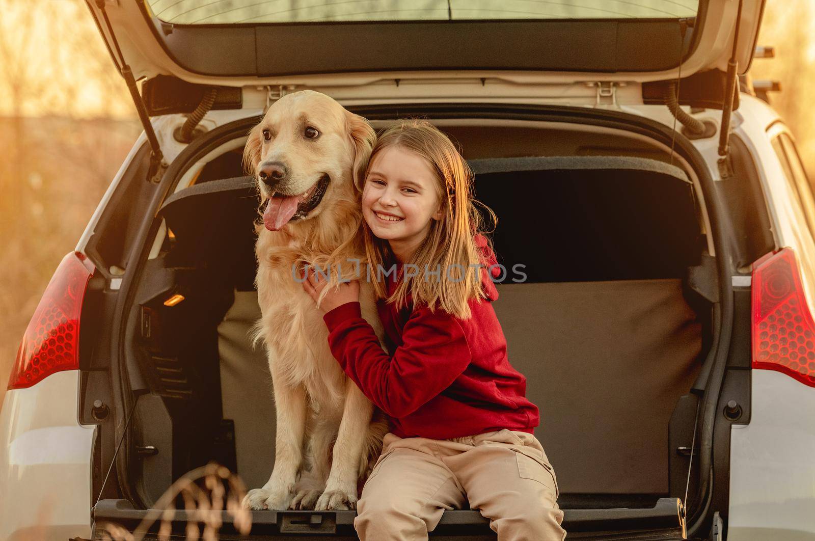 Girl child hugging golden retriever dog sitting in car trunk outdoors. Pretty kid petting doggy pet labrador and smiling in vehicle at nature