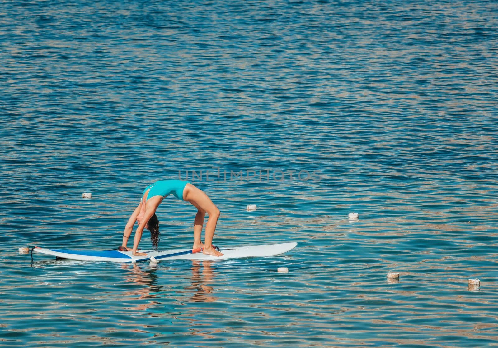 Young woman in blue swimsuite doing yoga on sup board with paddle. Balanced pose - concept of harmony with the nature, free and healthy living, freelance, remote business