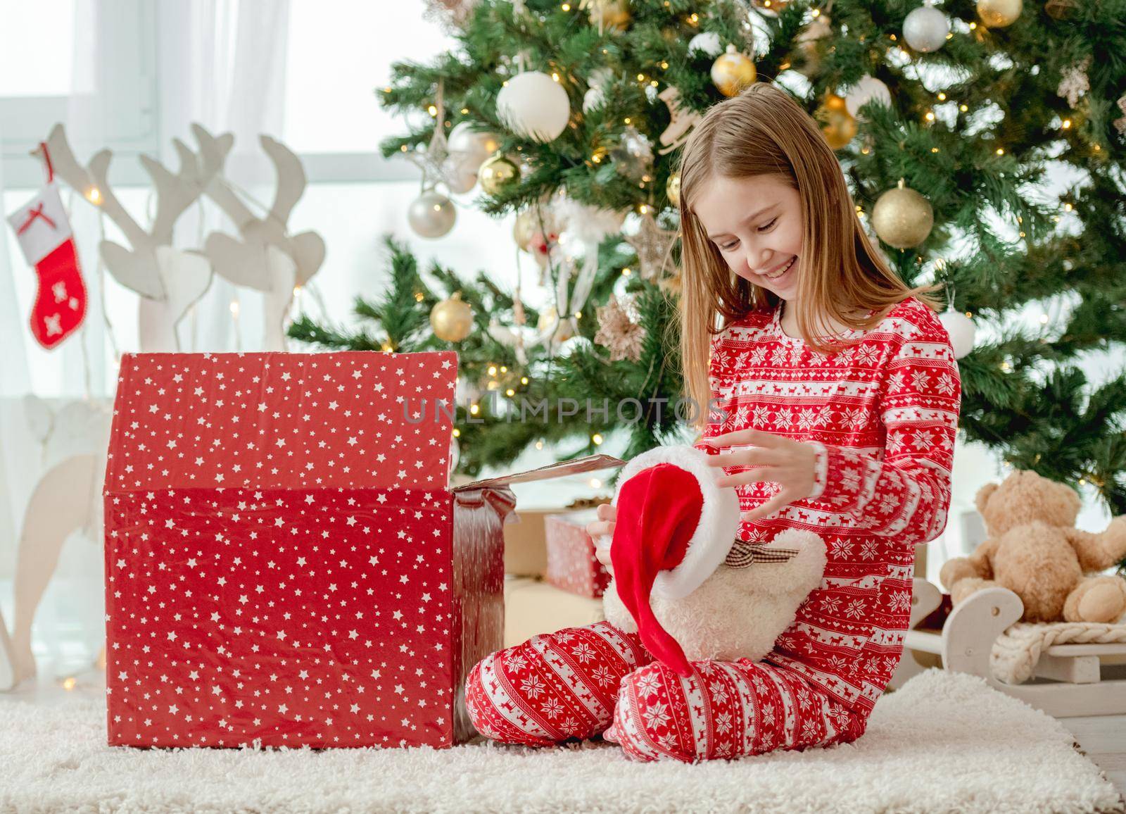 Pretty child girl open Christmas red box at home with traditional decorated tree and happy with gift toy teddy bear. Kid celebrating New Year with presents