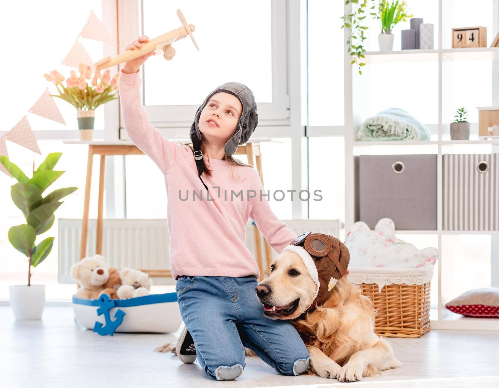 Little girl wearing pilot hat sitting on the floor with golden retriever dog in pilot glasses and play with wooden plane