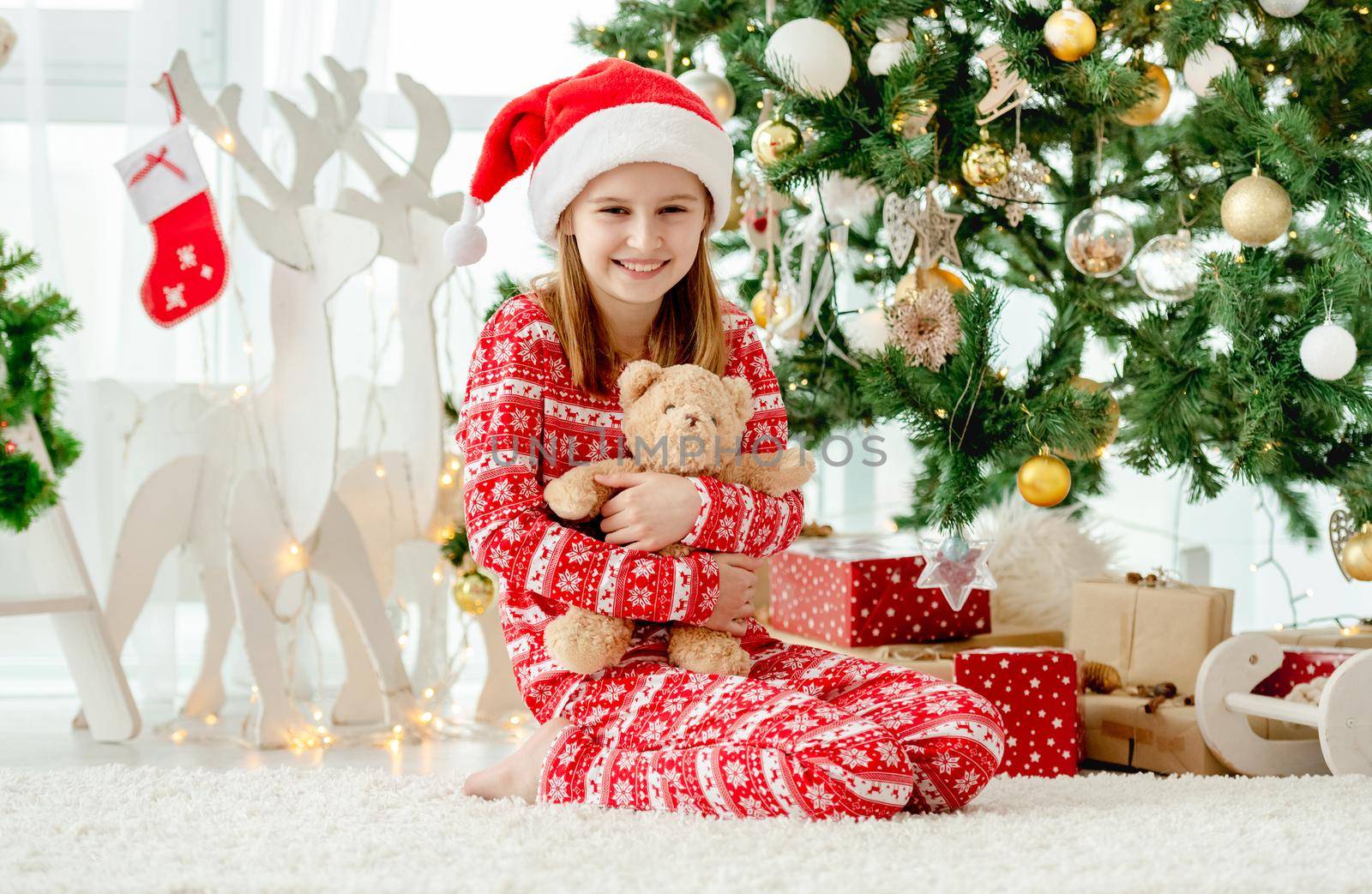 Happy child girl hugging teddy bear toy at home with Christmas tree on background. Cute kid with gifts at New Year time