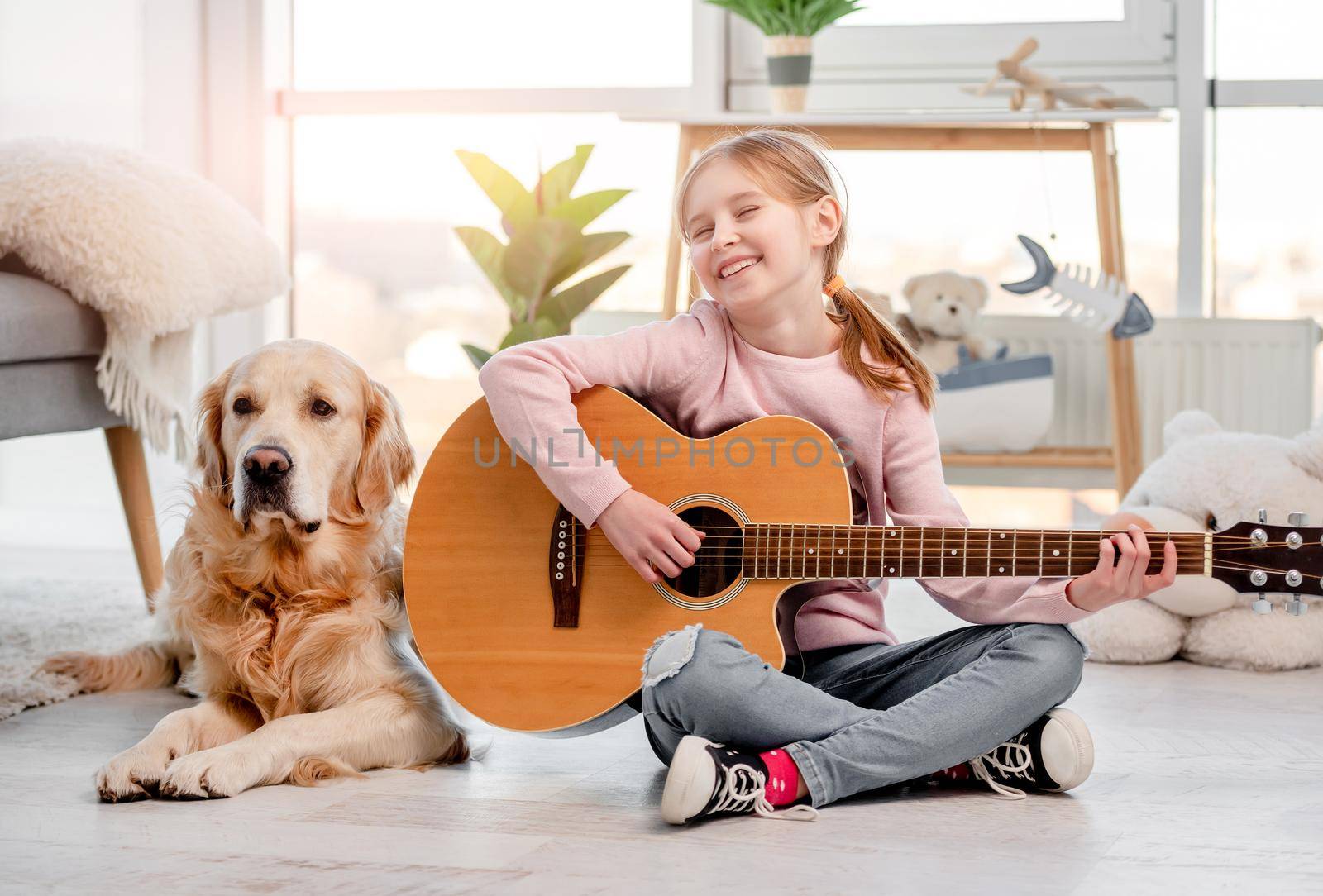 Little girl sitting on the floor and playing guitar with golden retriever dog lying next to her
