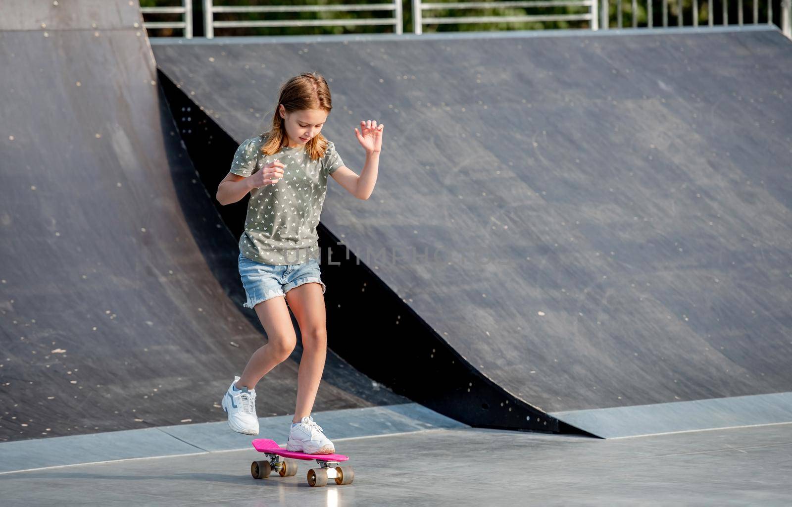 Preteen girl riding on skateboard at city park ramp. Beautiful female child with board outdoors at summer