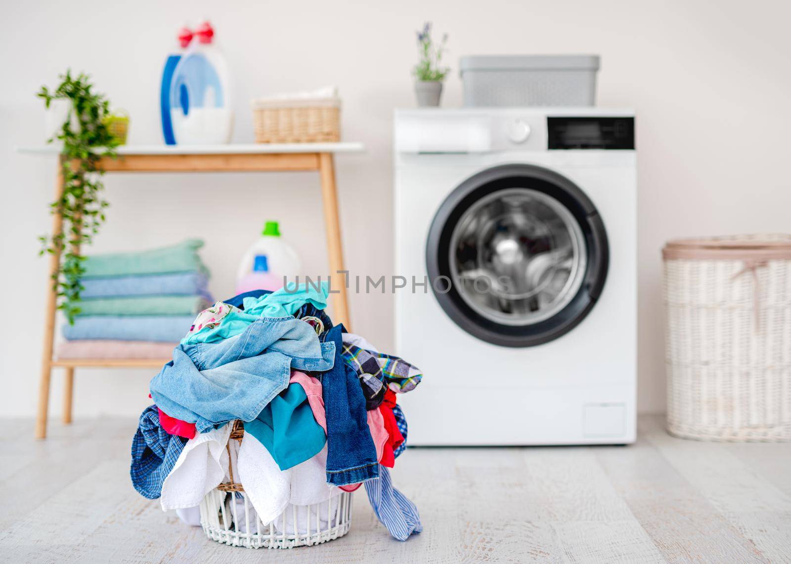 Heap of clothes for washing in basket standing on floor near washing machine in light bathroom