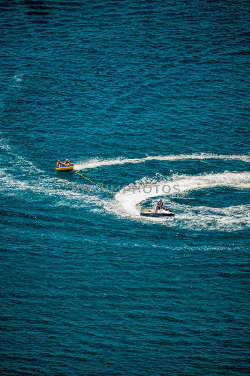 Happy people are going to swim on an air mattress behind a jet ski. Tourists ride the inflatable watercraft boat. Sea attraction. Jet ski skating people on a large Inflatable raft. by panophotograph
