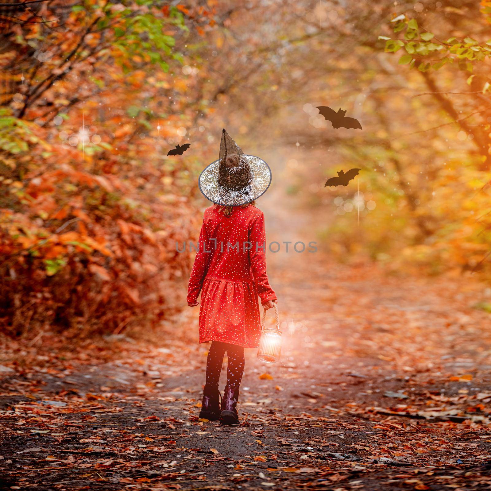Little girl in witch hat holding lantern while standing backwards in autumn wood with flying bats