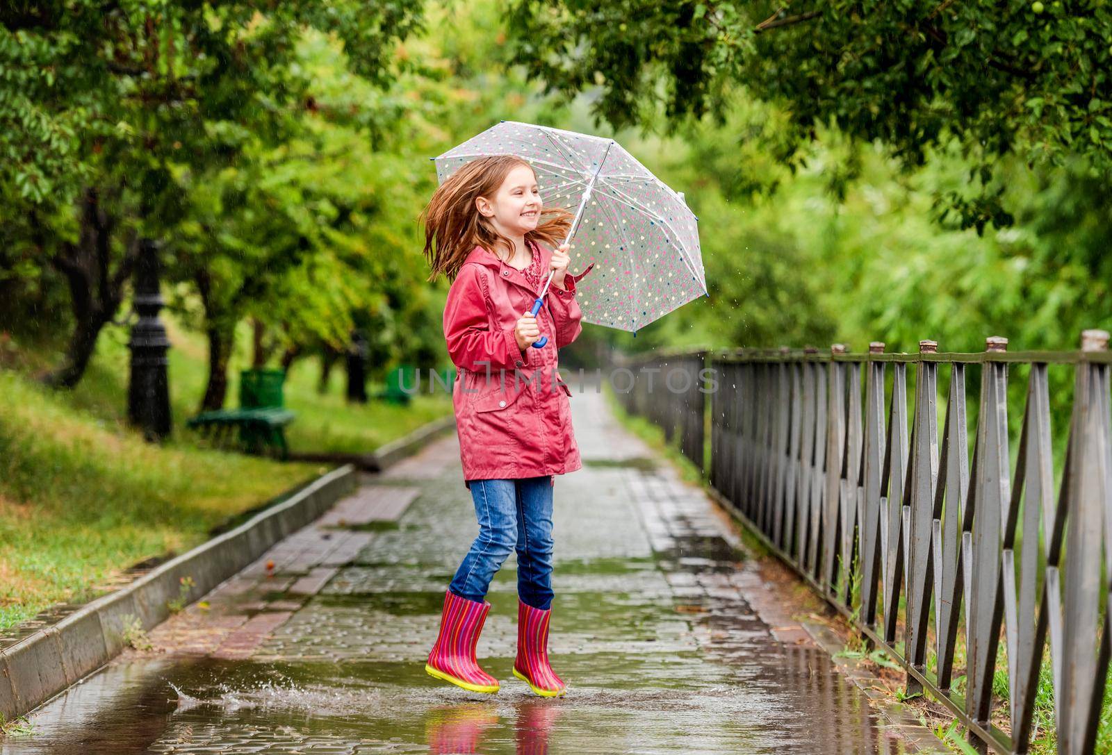 Little girl with umbrella jumping in puddle during rainy walk in park