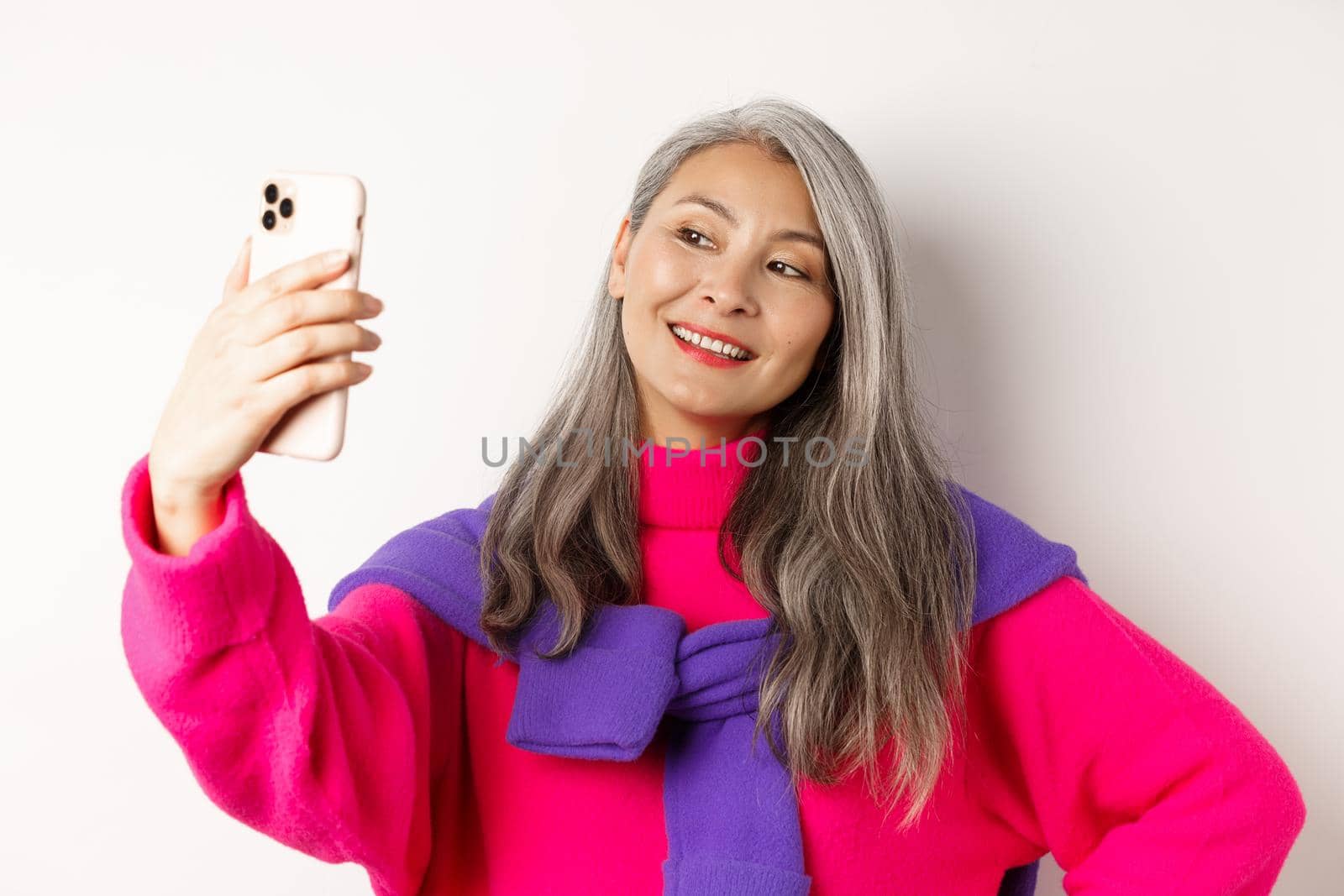 Beautiful and stylish asian senior woman taking selfie on smartphone, smiling and posing in trendy pink sweater, standing over white background.