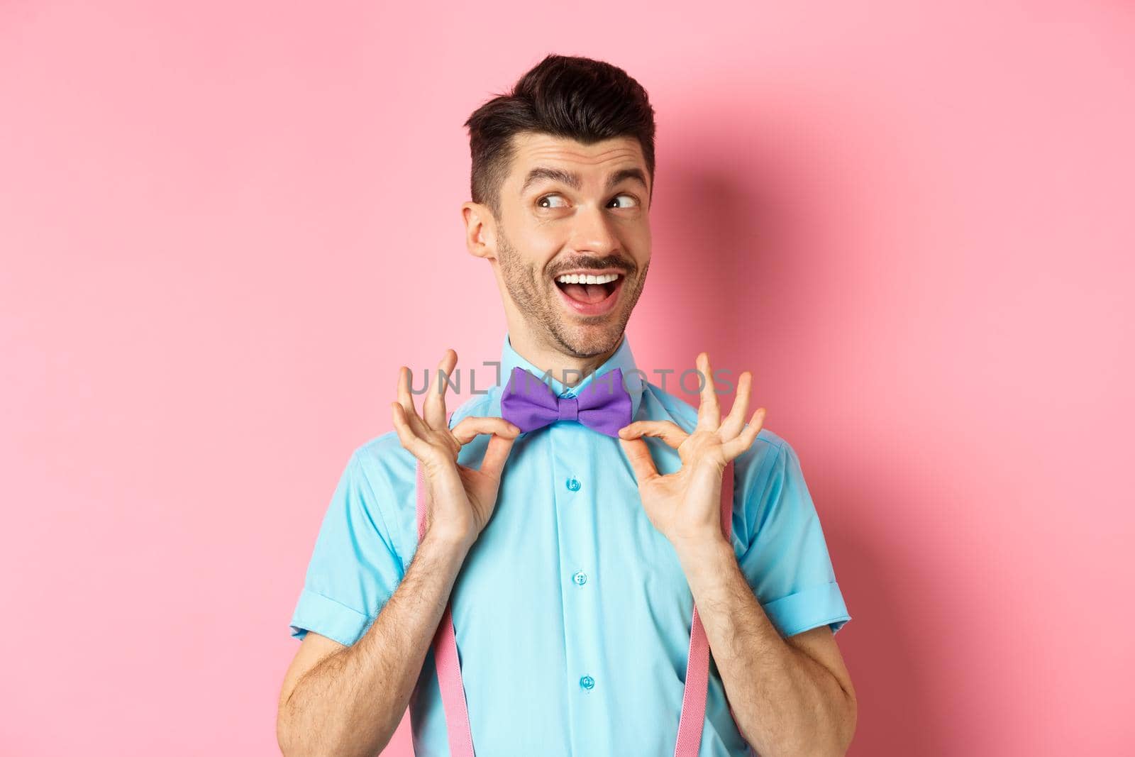 Cheerful and confident young man with moustache, fixing bow-tie on neck and looking left pensive, smiling happy, standing on pink background.