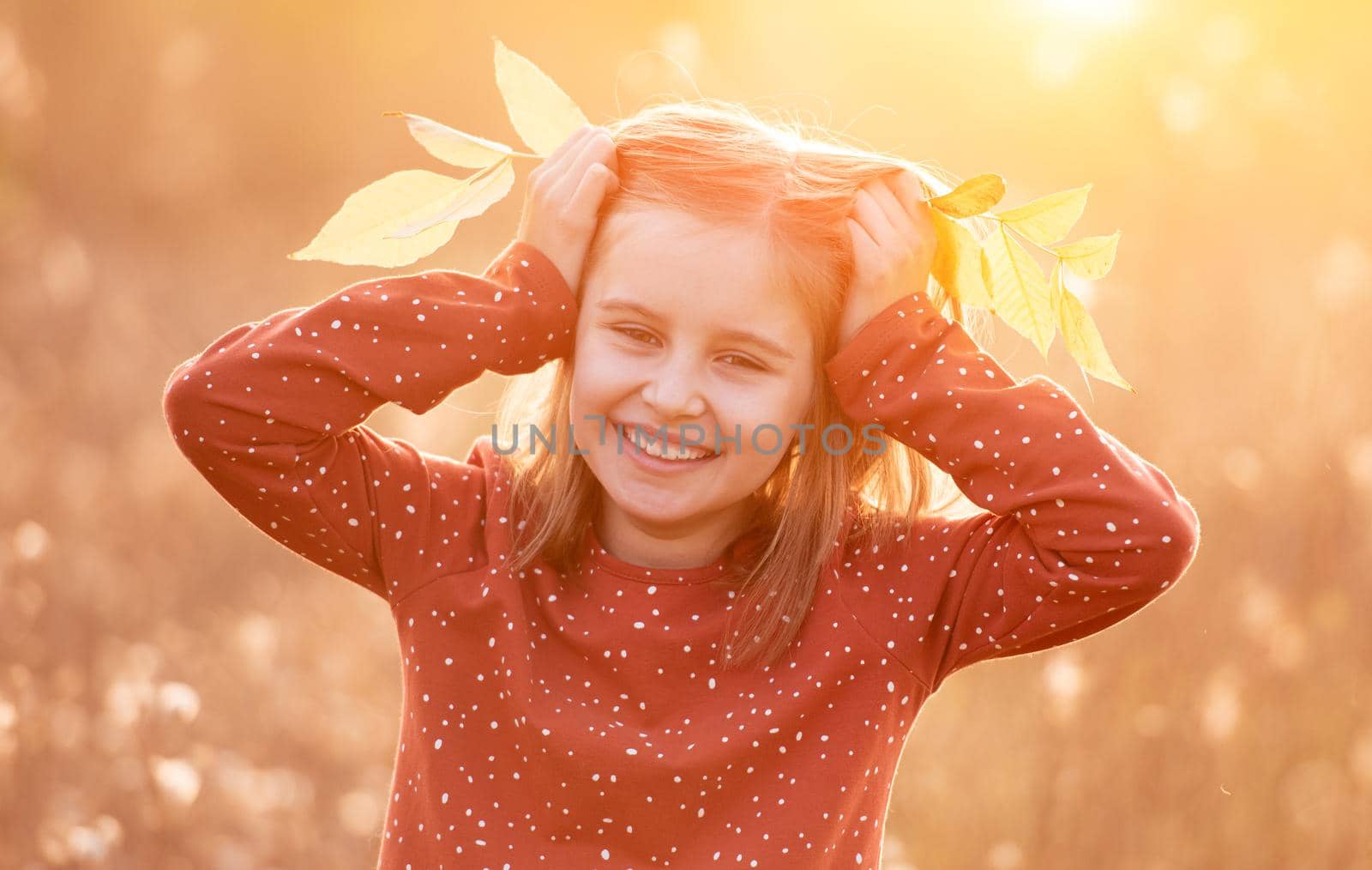 Portrait of smiling little girl holding autumn leaves on head in sunlight