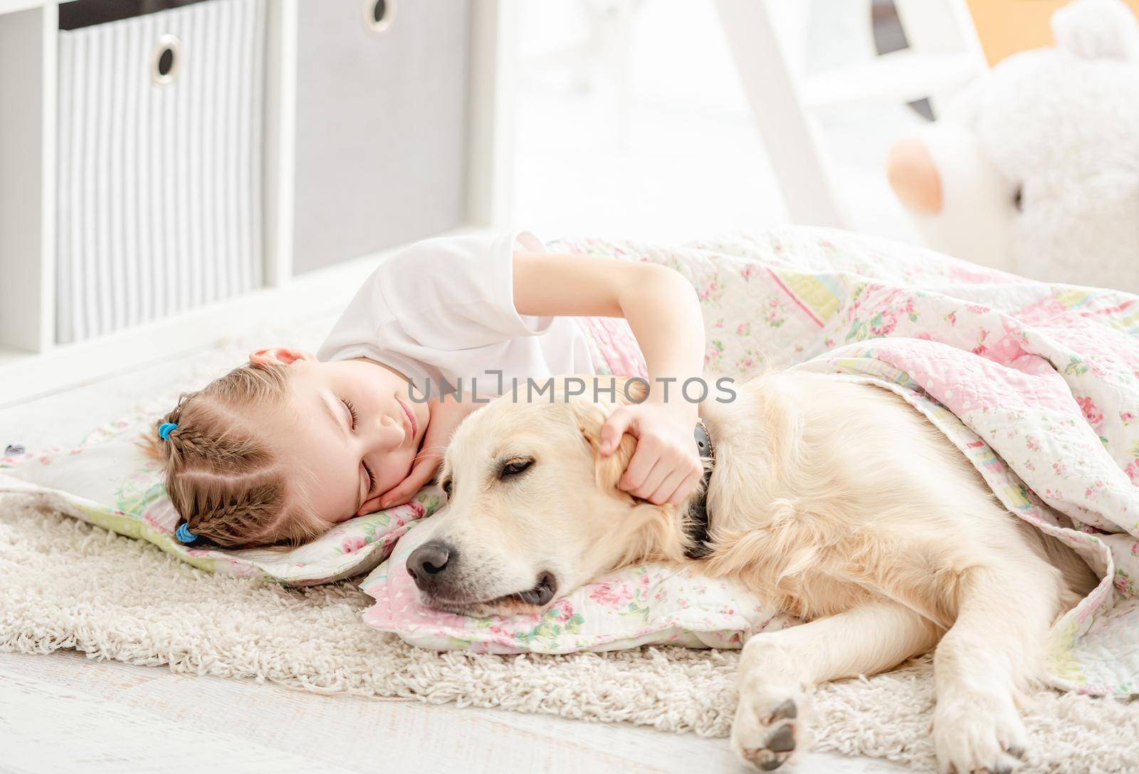 Happy little girl sleeping with lovely dog under blanket on floor in light room