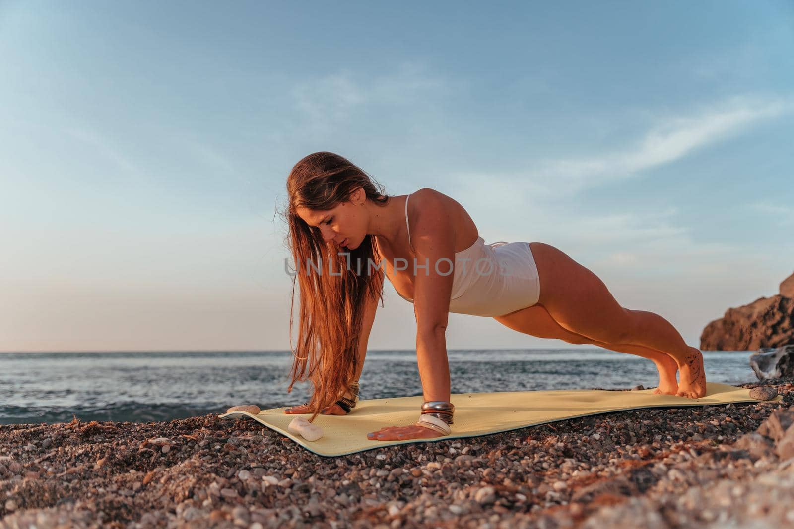 Young woman with long hair in white swimsuit and boho style braclets practicing outdoors on yoga mat by the sea on a sunset. Women's yoga fitness routine. Healthy lifestyle, harmony and meditation by panophotograph