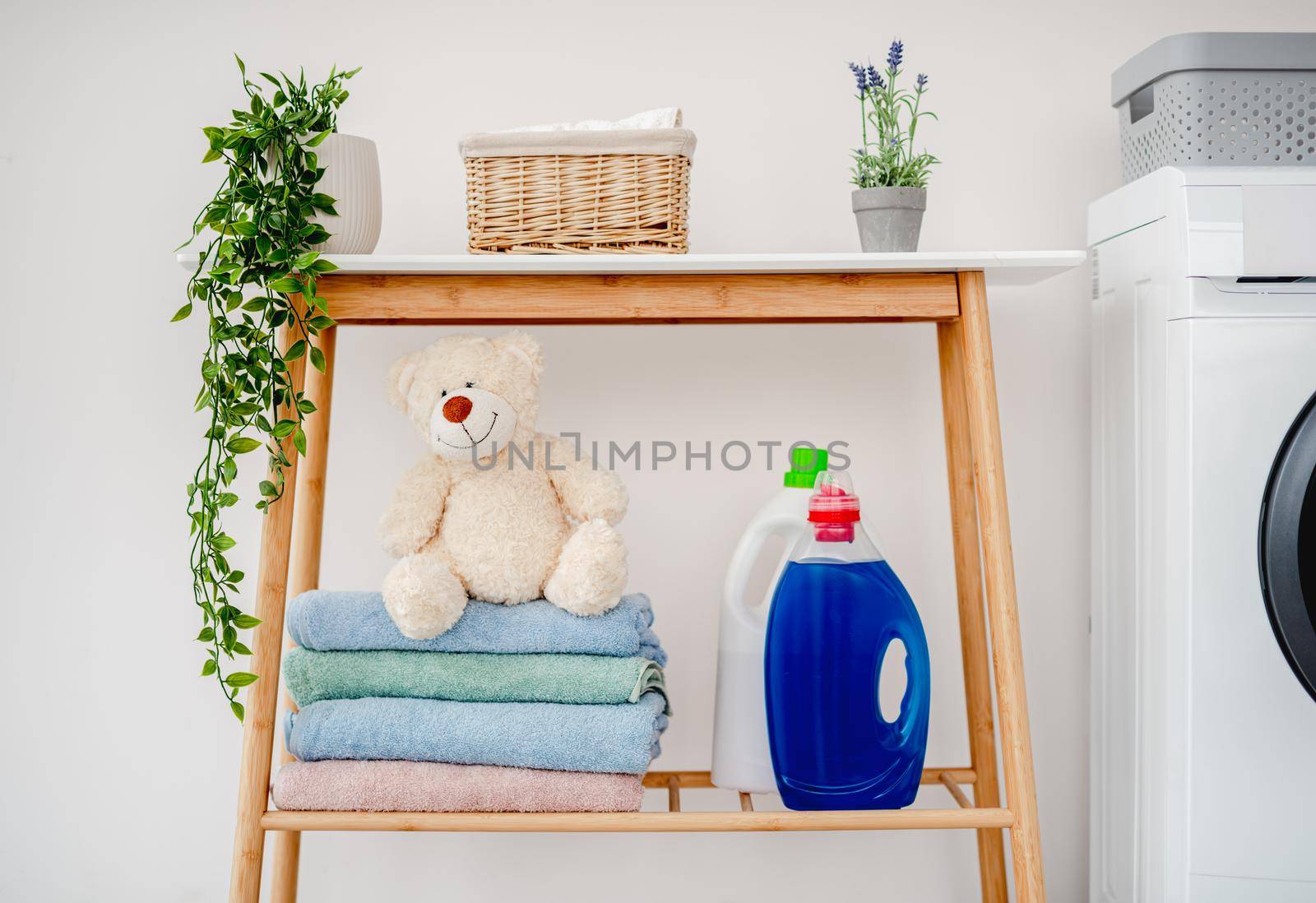 Shelf with washing powders, clean towels and plush teddy standing near washing machine in light room