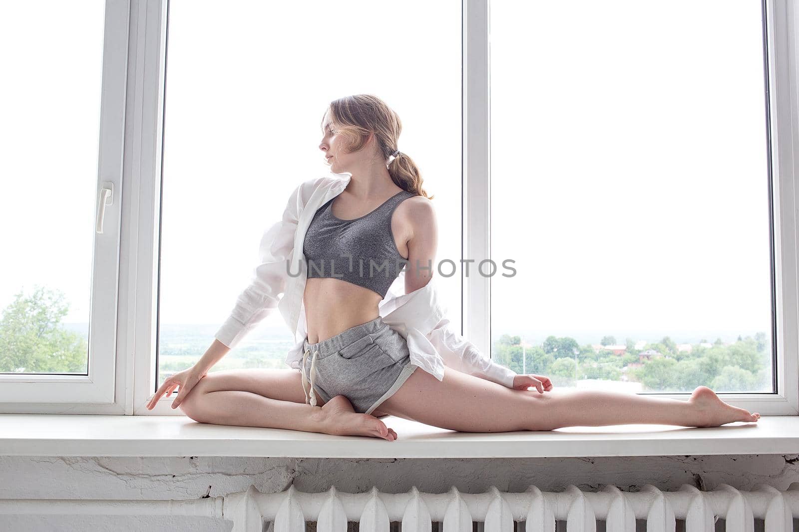 girl practicing yoga, doing splits, stretching exercise on windowsill at home by artemzatsepilin