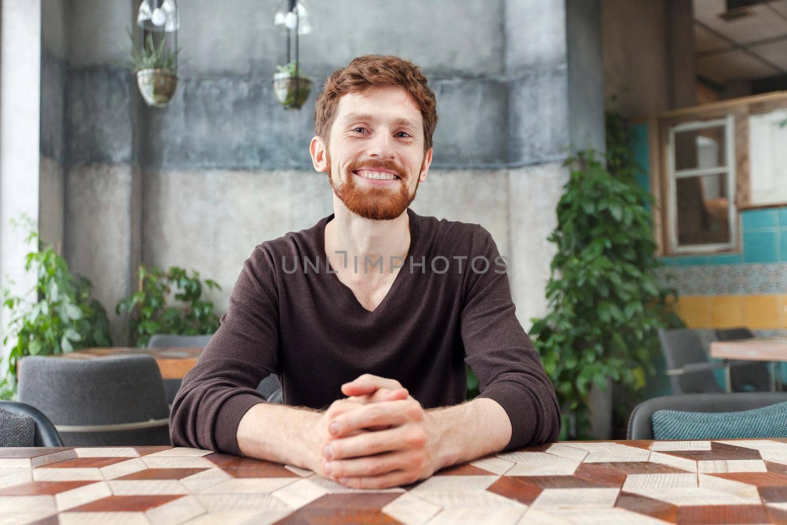 Happy adult bearded redhead man sitting at table in coffee shop and smiling at camera
