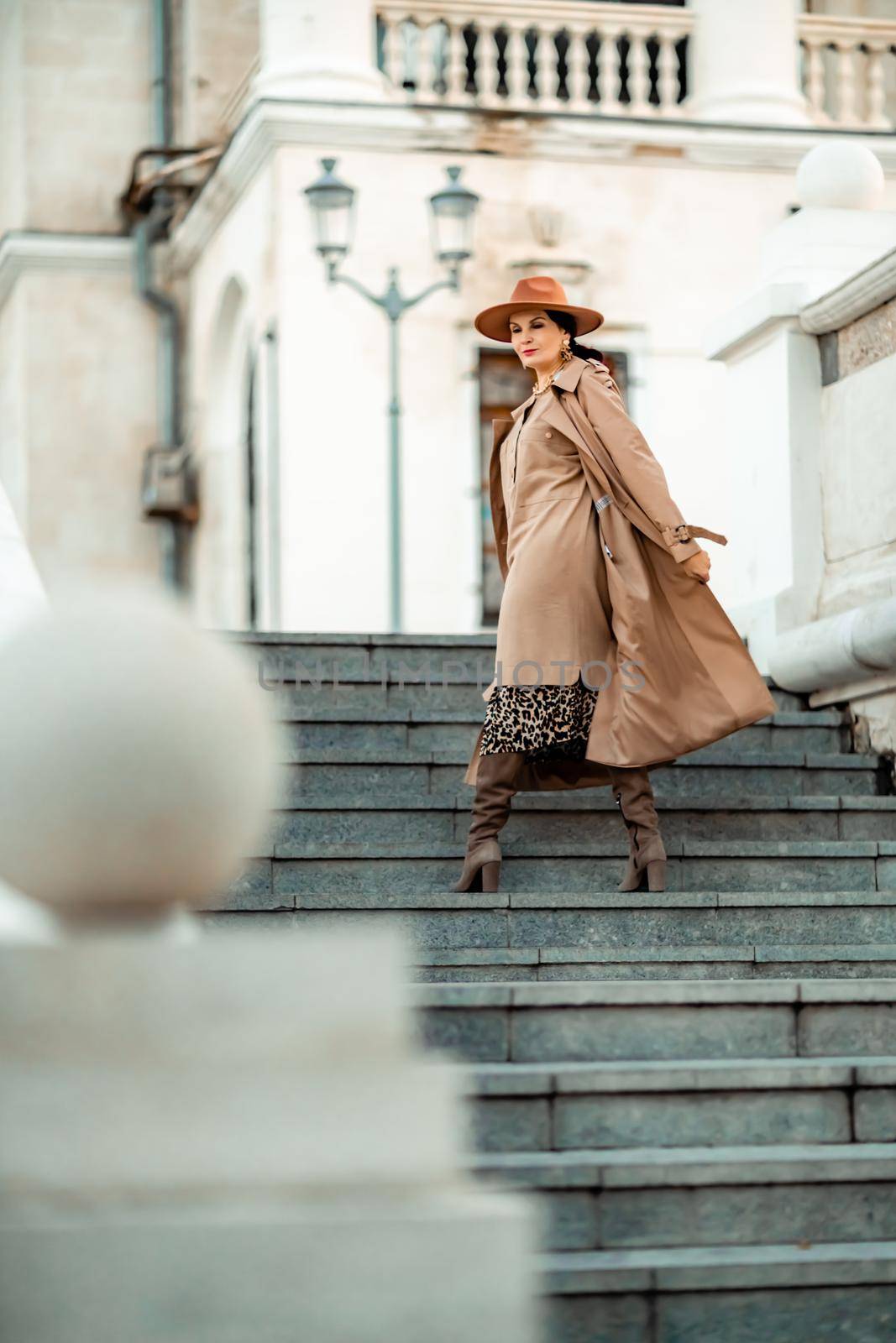 Outdoor fashion portrait of young elegant fashionable brunette woman, model in stylish hat, choker and light raincoat posing at sunset in European city