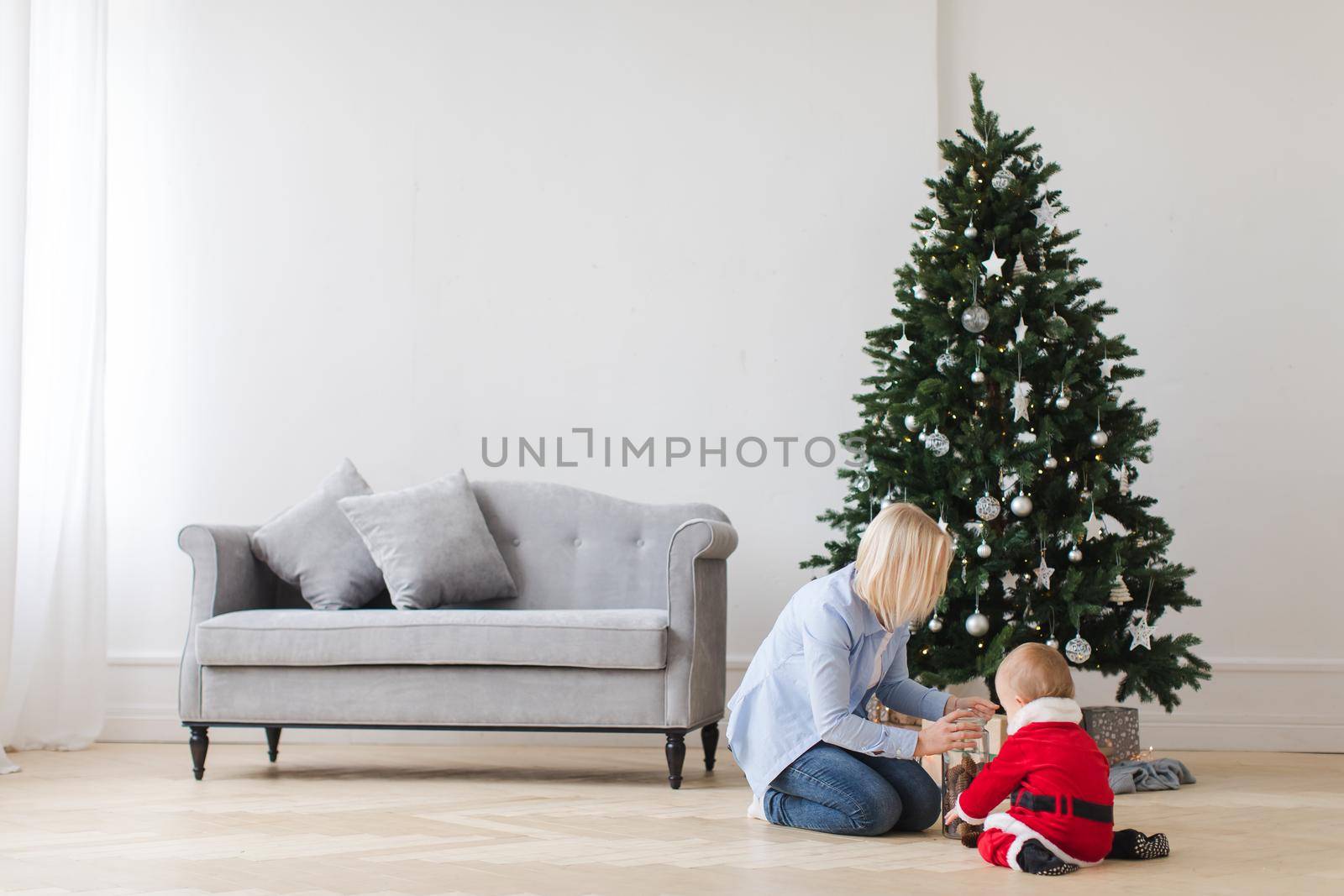 Woman with adorable child in Santa costume sitting near Christmas tree and exploring presents.