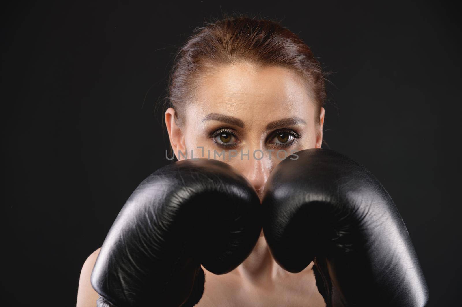 Young caucasian sportswoman in sportswear and large black boxing gloves looks into the camera and covers her face with gloves. Copy space.