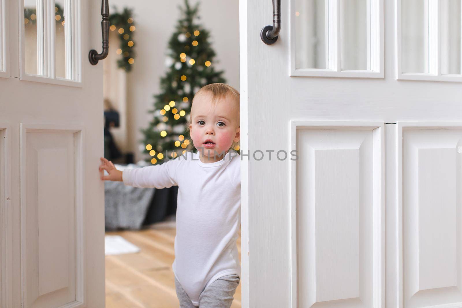 Adorable infant standing in doors looking at camera on background of Christmas tree in bedroom.