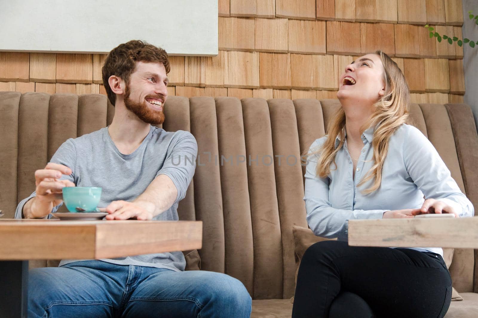 Cheerful man and woman sitting at table in modern coffee shop with mugs and laughing happily