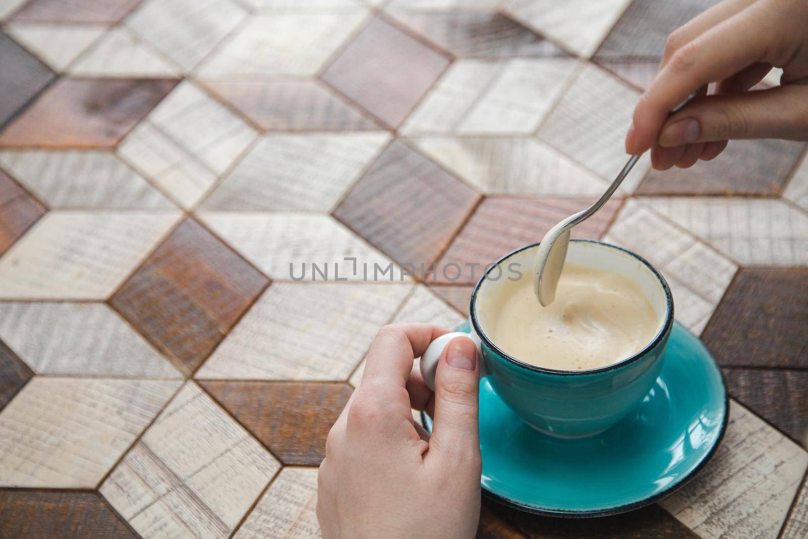 Cup of cappuccino with milk foam on wooden table. Female hand holds spoon with sugar