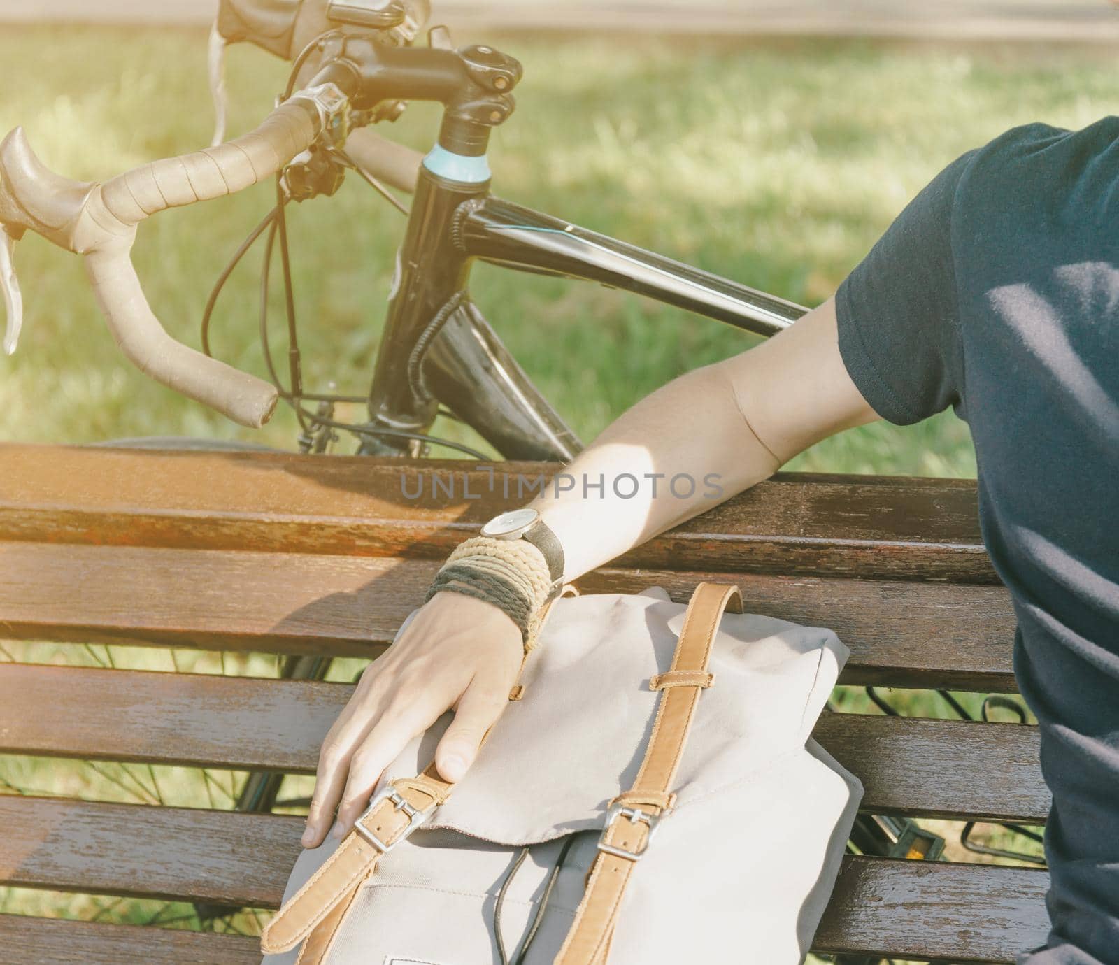 Unrecognizable young man with backpack and bicycle rests on wooden bench in summer park.