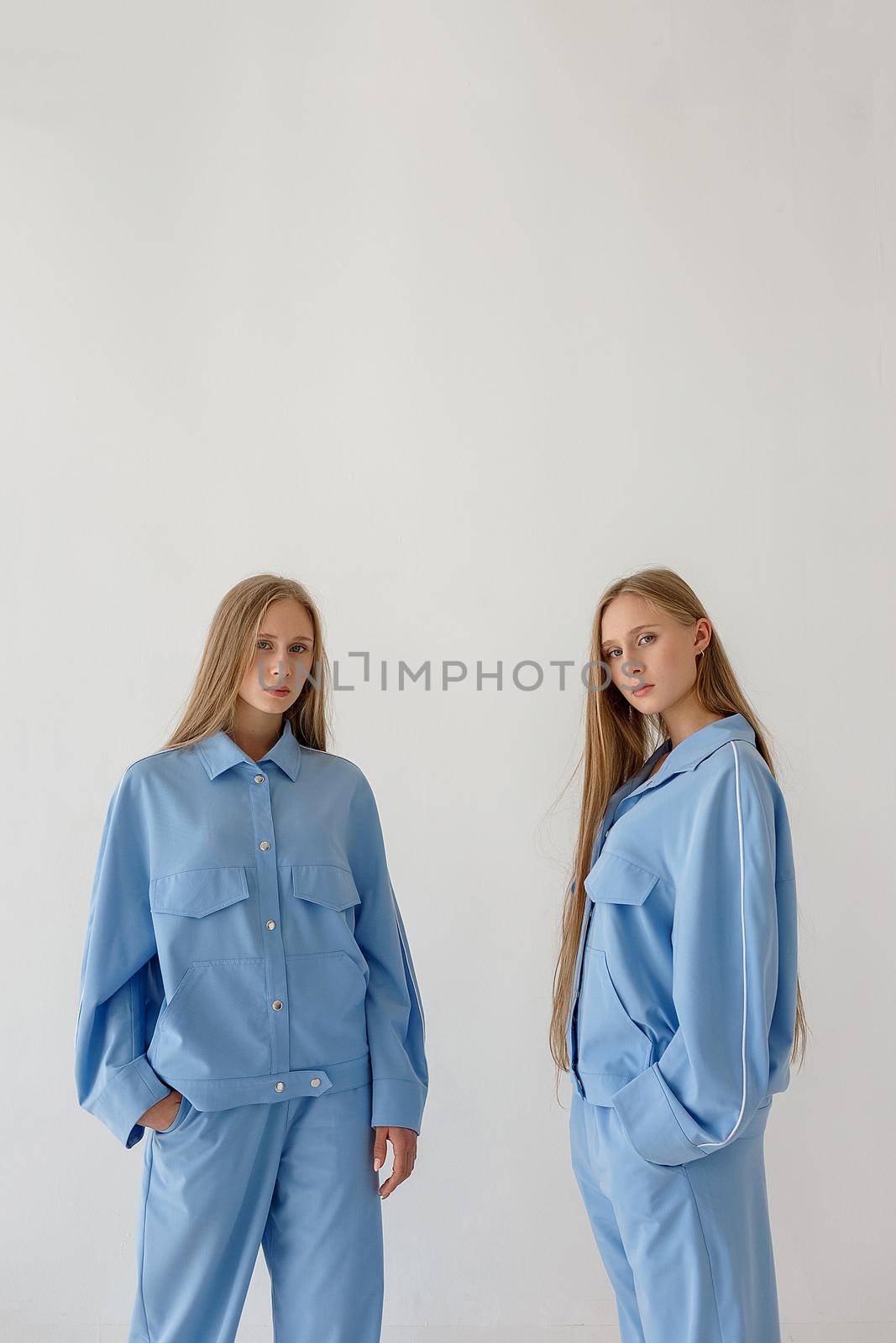 two young twin sisters with long blond hair posing on white background in oversize clothes with bare feet and looking at camera. They wear blue suits that look like pajamas. Stylish fashion photoshoot