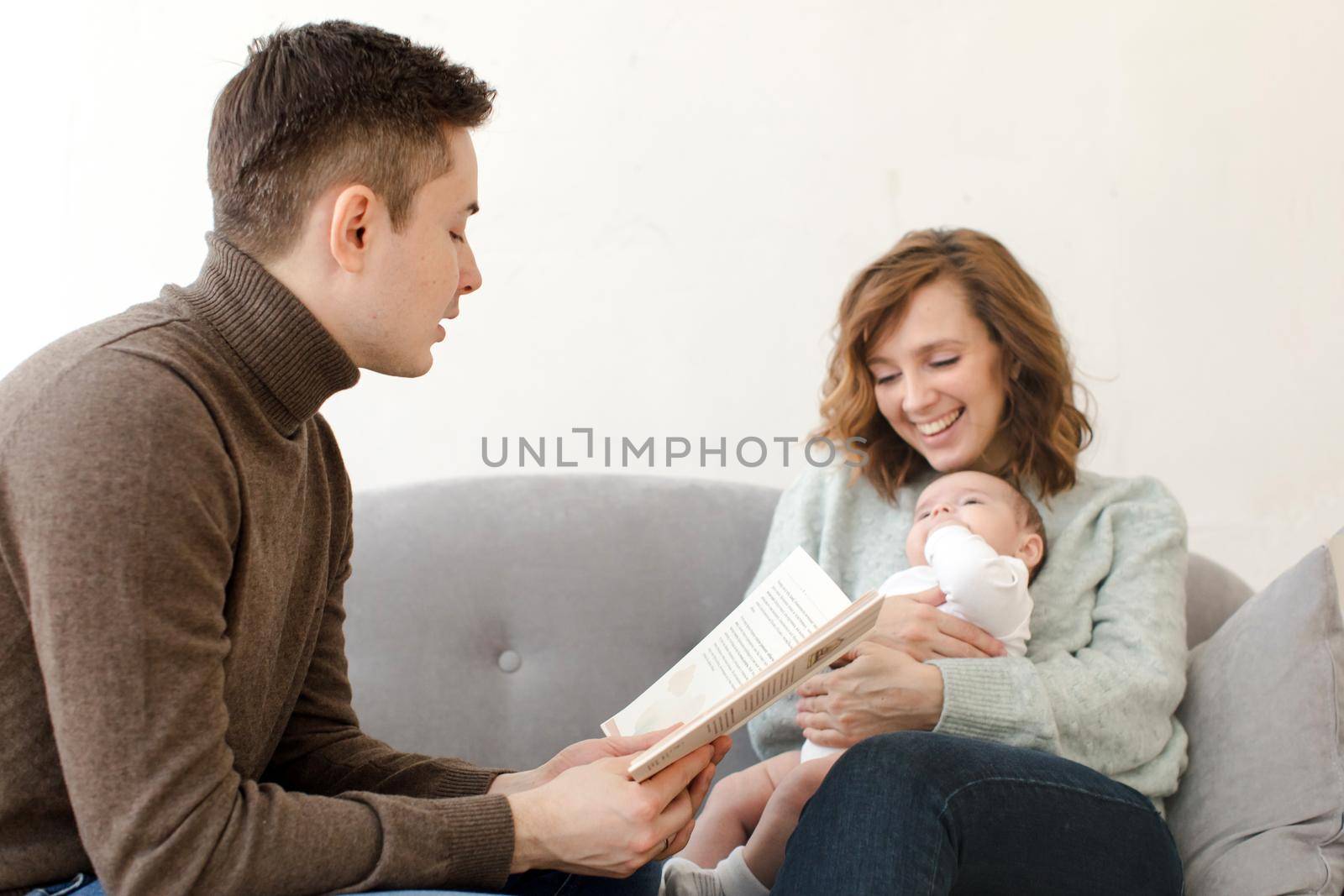 Young father reading book for little child sitting on knees of young mother in living room